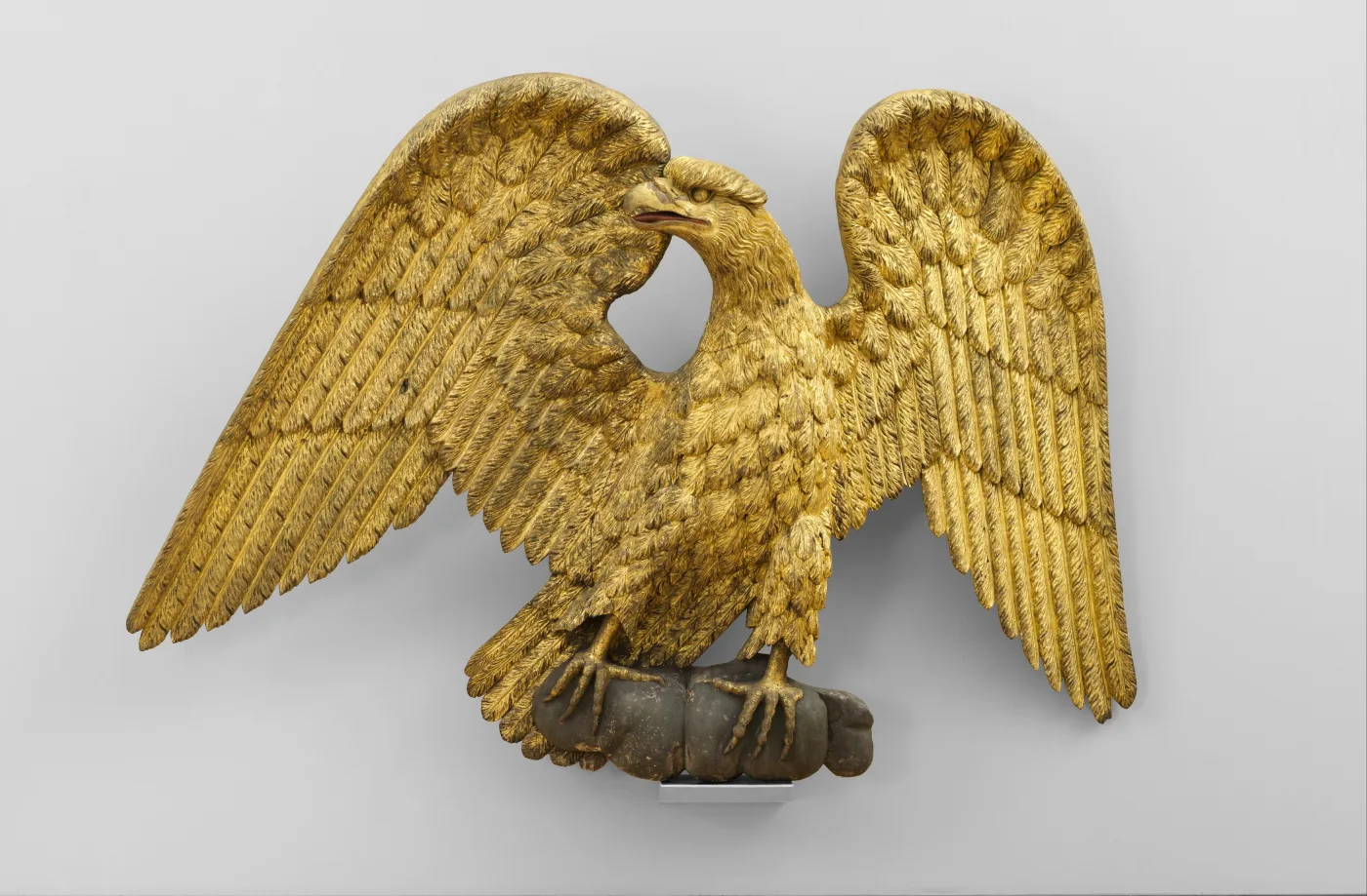 Gilded wooden eagle with both wings spread wide and level, its head turned sharply to one side, standing on a plain base