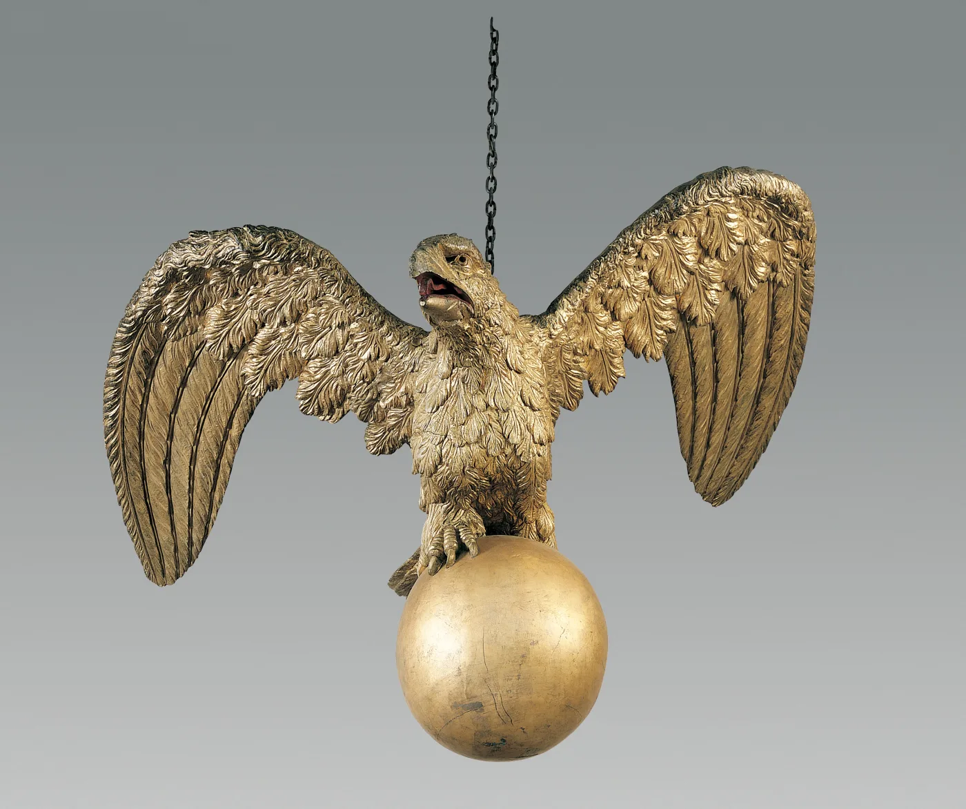 Gilded carved wooden eagle with wings raised and head turned, gripping a gilt sphere, suspended from a chain against a plain grey ground