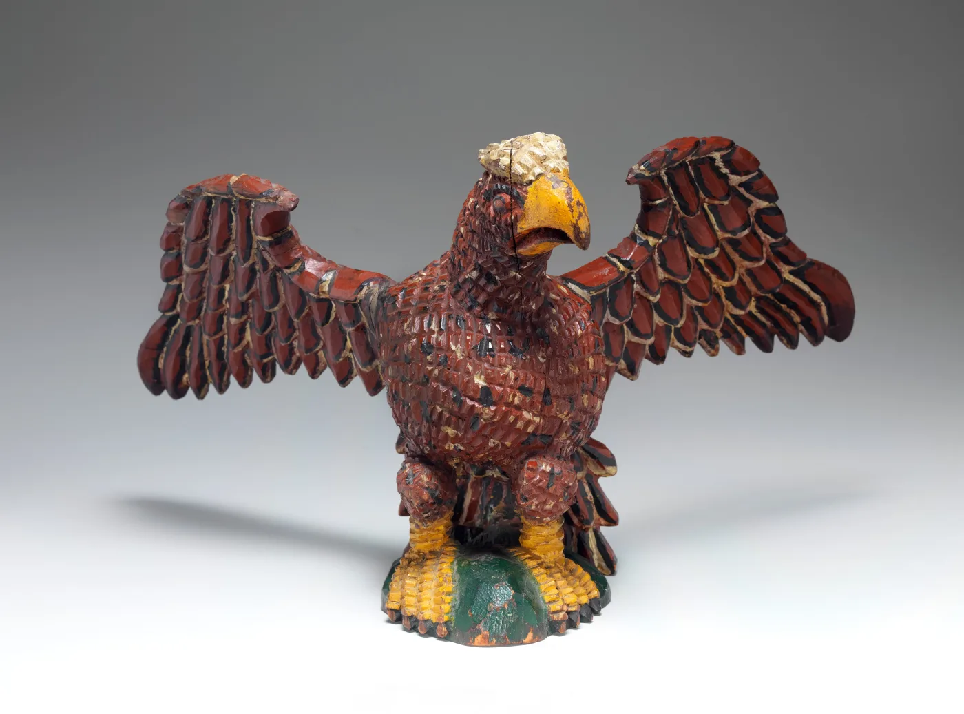 Small carved eagle painted in red-brown with a yellow beak, its wings notched into feather shapes and its body upright on a green mound