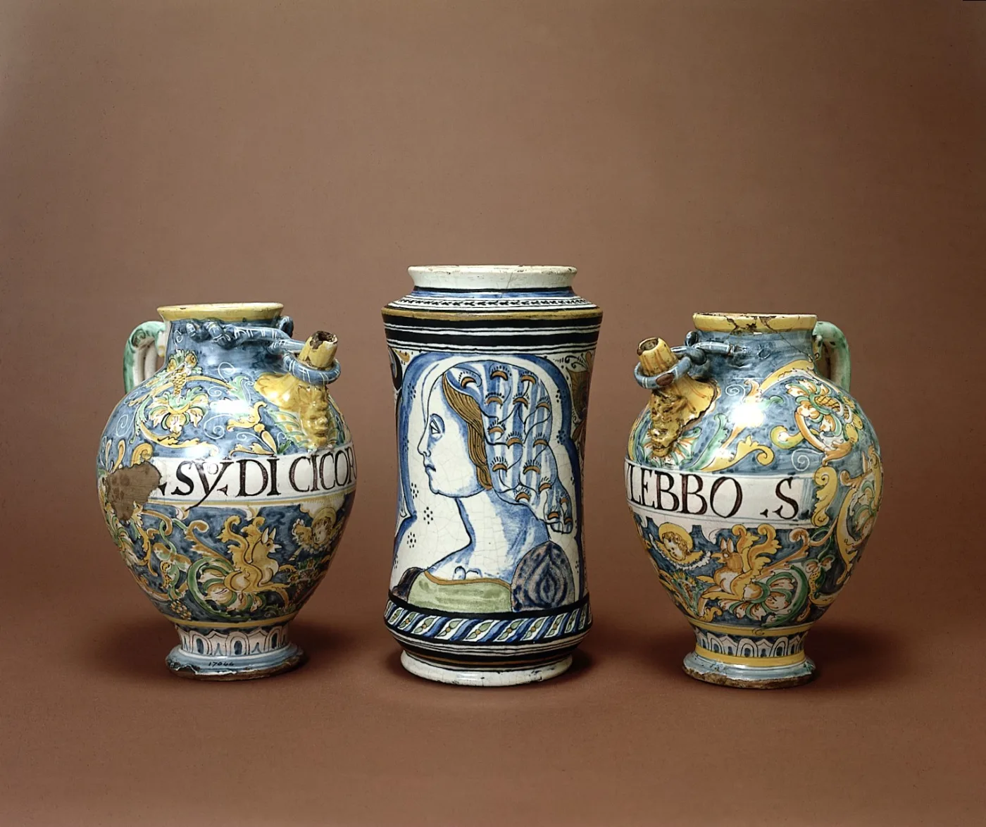 Three ceramic pharmacy jars: two ovoid handled jars painted with drug names in gothic blackletter on white bands, flanking a taller waisted cylindrical albarello painted with a woman's head in profile