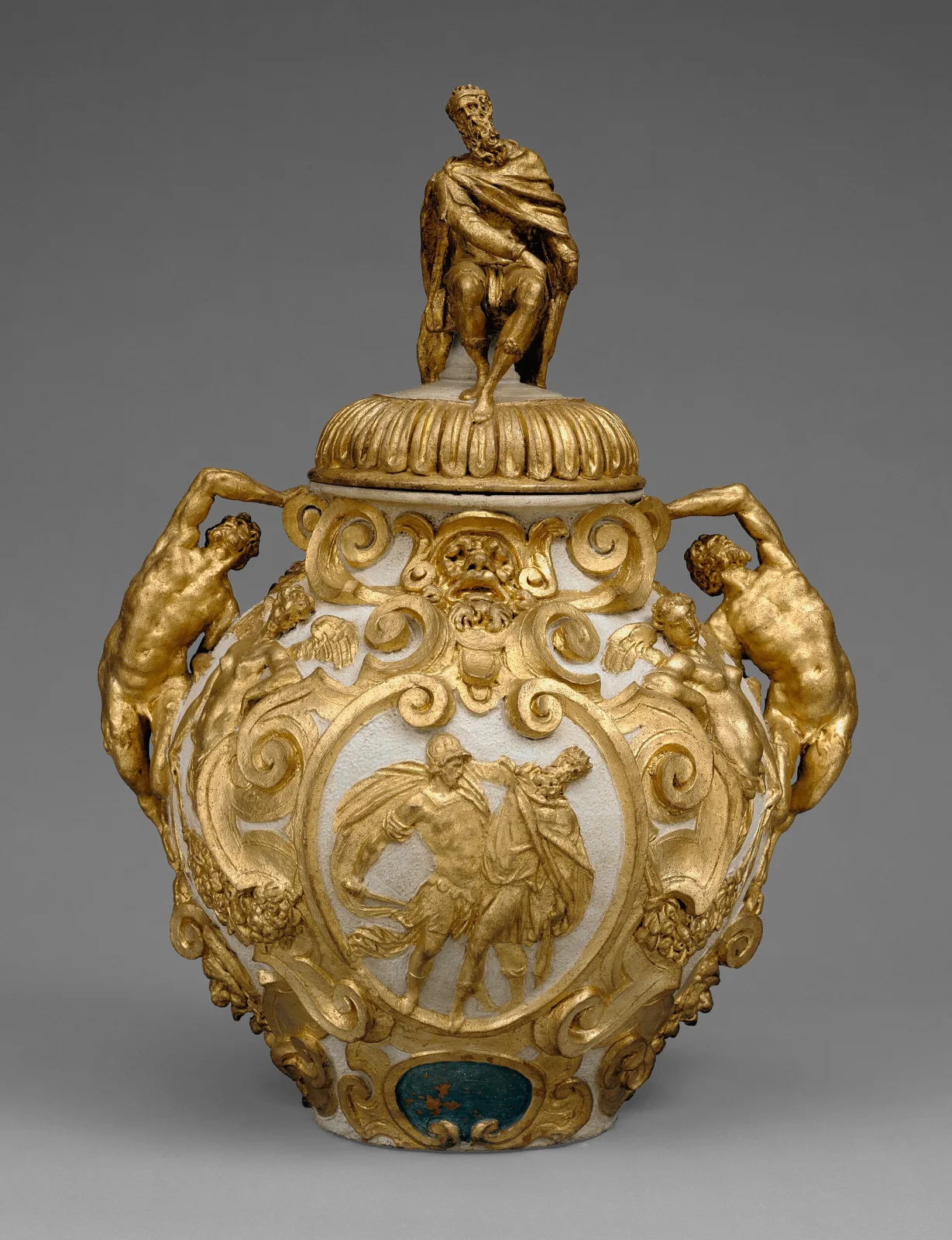 Elaborate lidded drug jar with gilded relief decoration, muscular figures forming the handles, a figural scene in an oval cartouche, and a seated gilded figure as the lid finial
