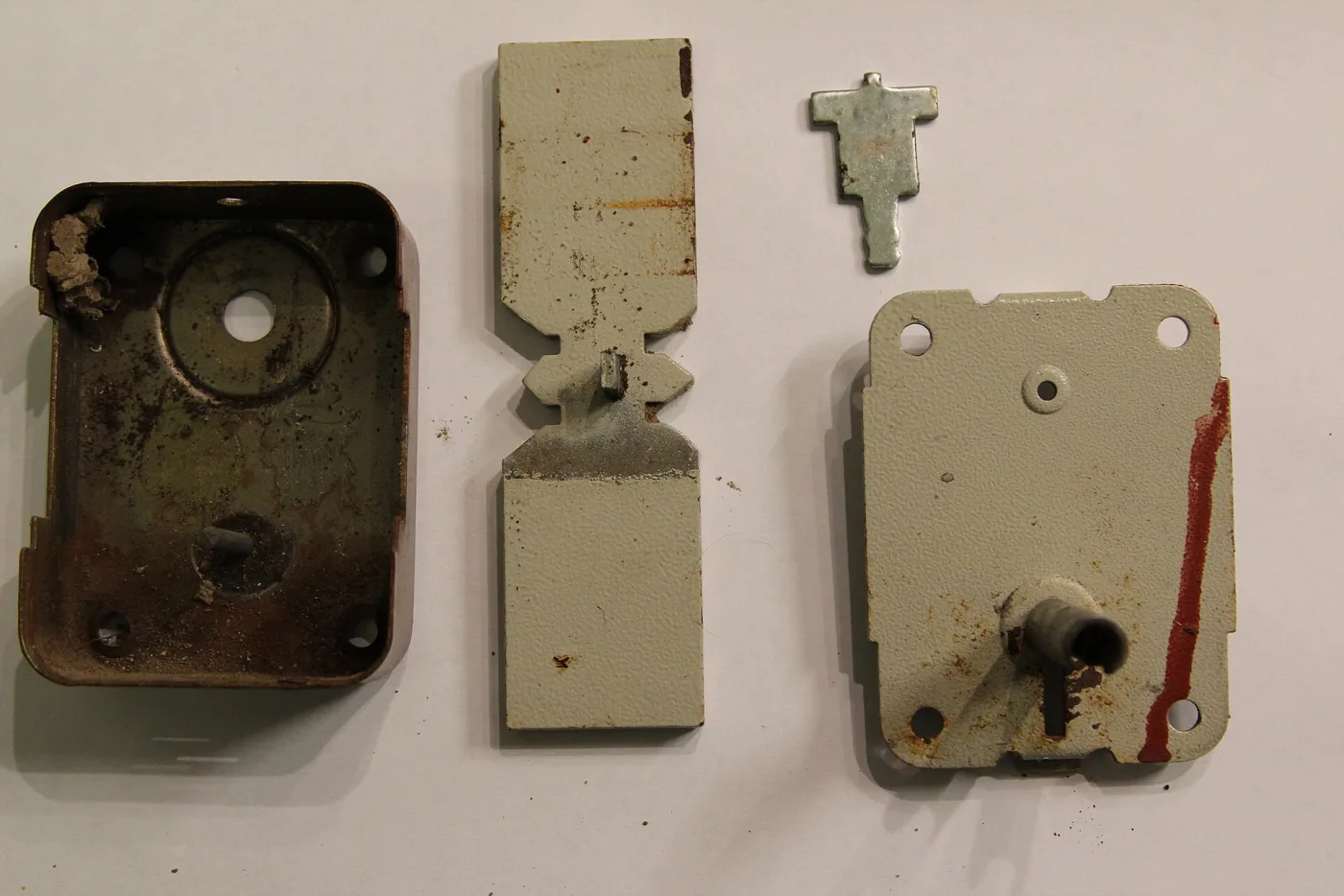 A rim lock taken apart: the iron case, a flat ward plate and the bolt laid out side by side on a white surface