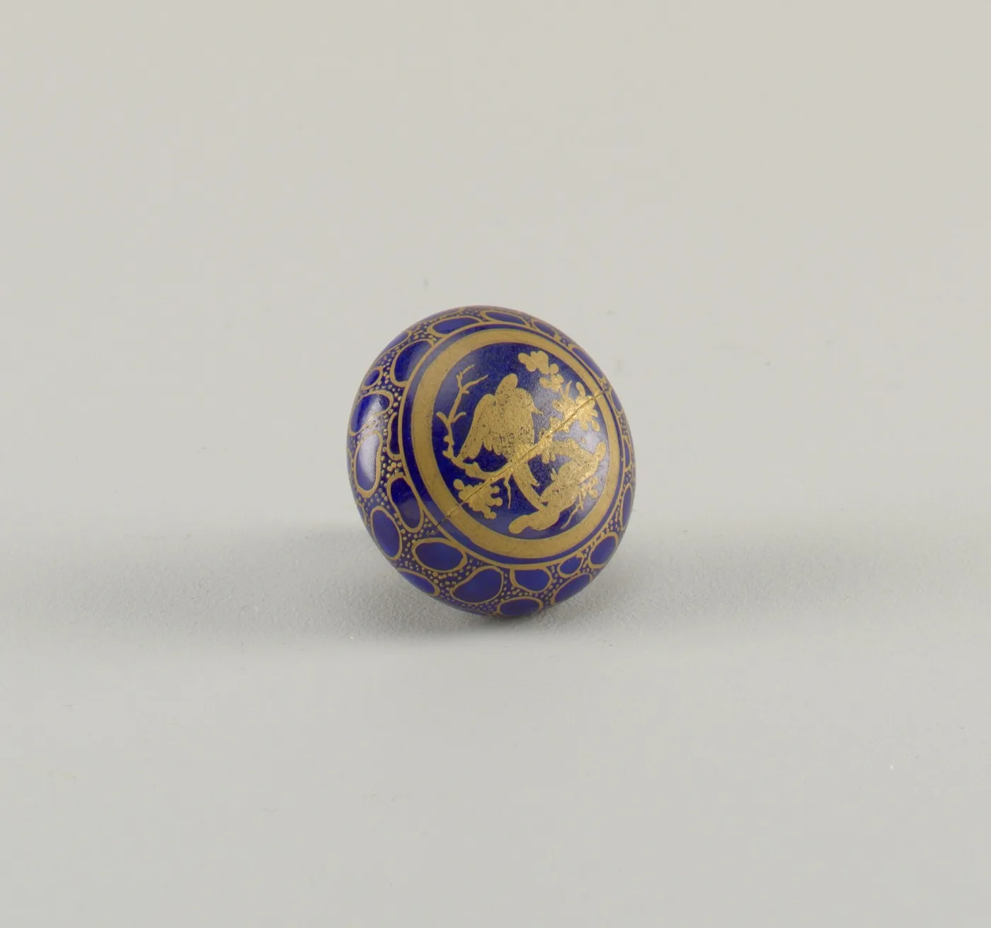 Small domed door knob in deep blue glass decorated with gilt foliage, photographed against a plain grey ground