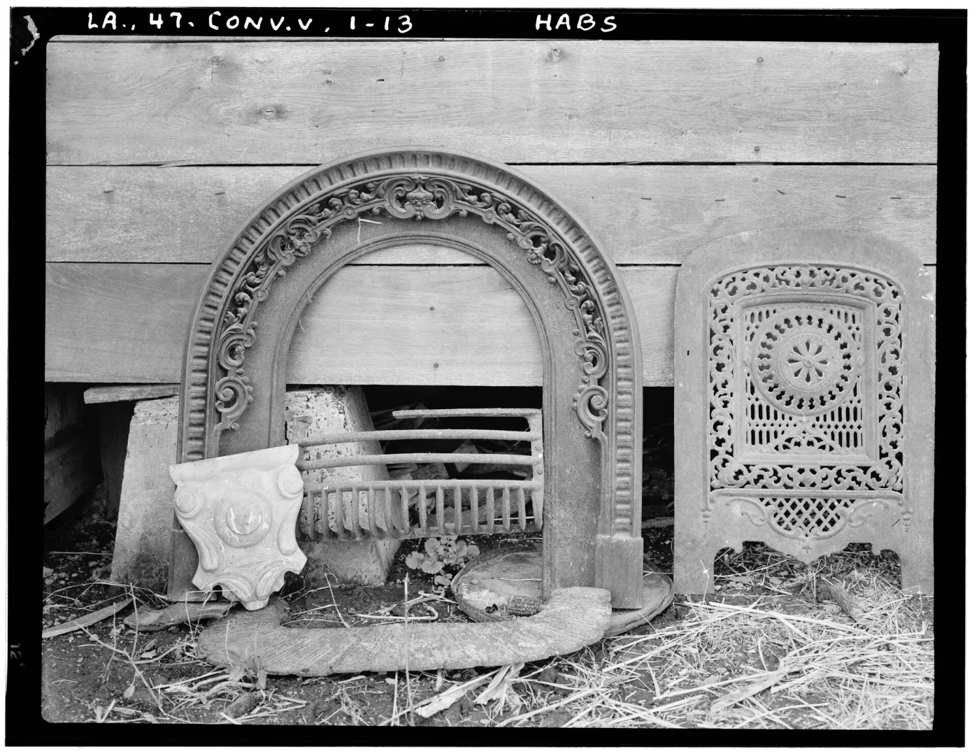 Black and white survey photograph of a cast iron fireplace arch and a pierced summer cover standing in straw against a plank wall