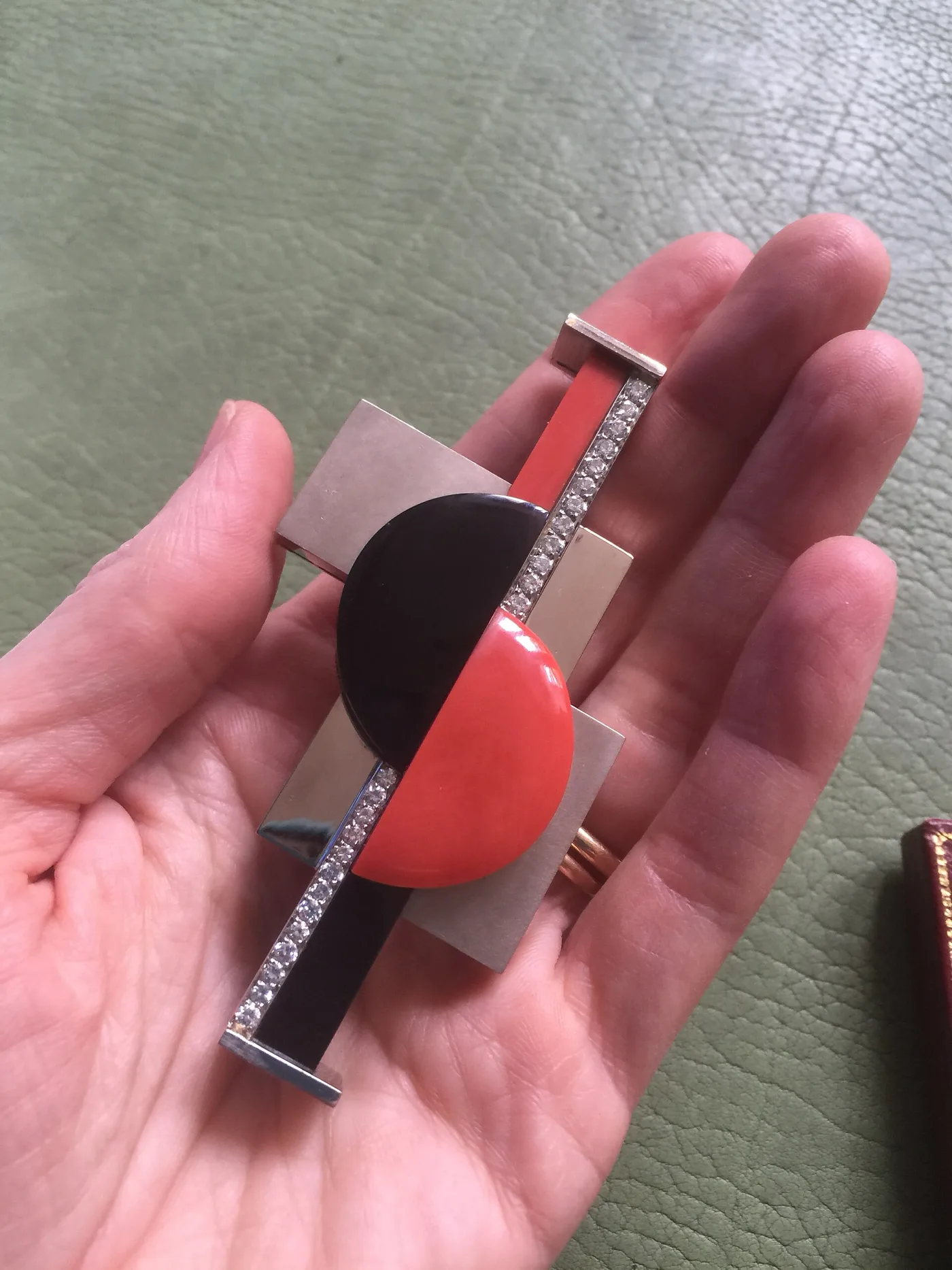 Geometric brooch held in a hand: polished white metal rectangles crossed by a diamond-set bar, with a black onyx disc overlapping a coral disc