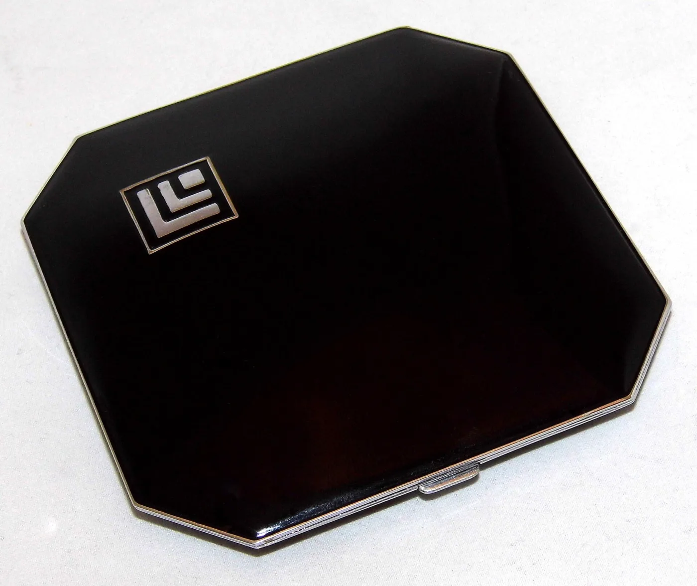 Octagonal powder compact with clipped corners, black lacquer lid, chrome rim and a white enamel monogram set in a small rectangle