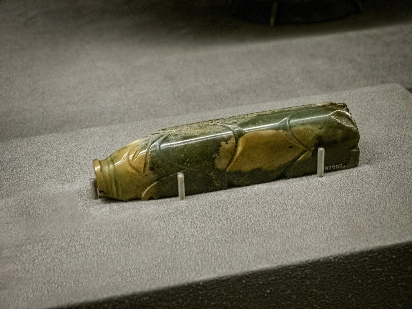 Elongated cylindrical jade carving of a recumbent pig, its form reduced to a few broad facets, resting on museum mounts under directed light