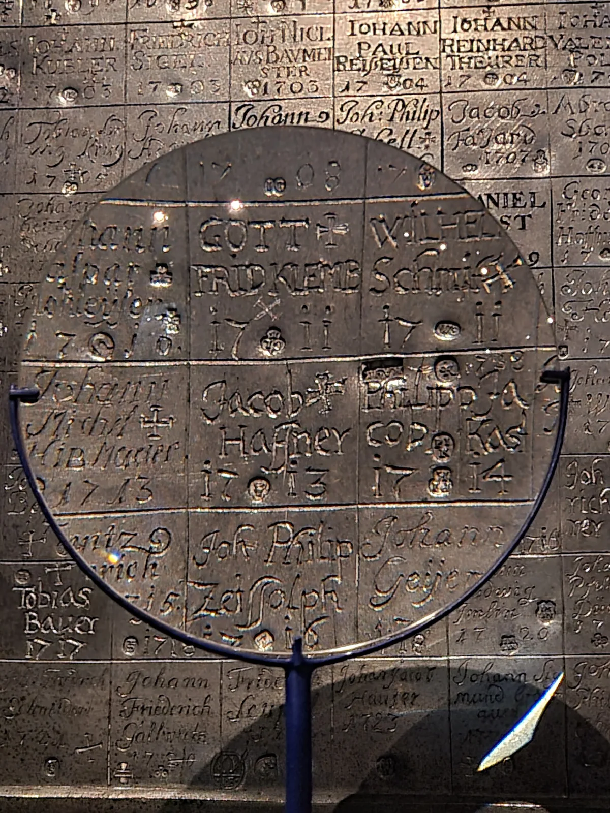Pewter plate covered in a grid of small struck punch marks with handwritten names and dates inked beside each one, displayed in a museum case