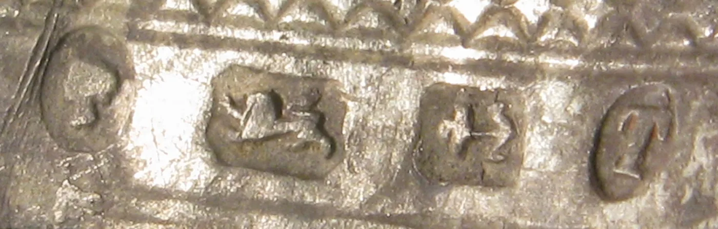 Extreme close-up along the side of a crushed silver thimble, showing an oval cartouche and a line of tiny struck marks beside it