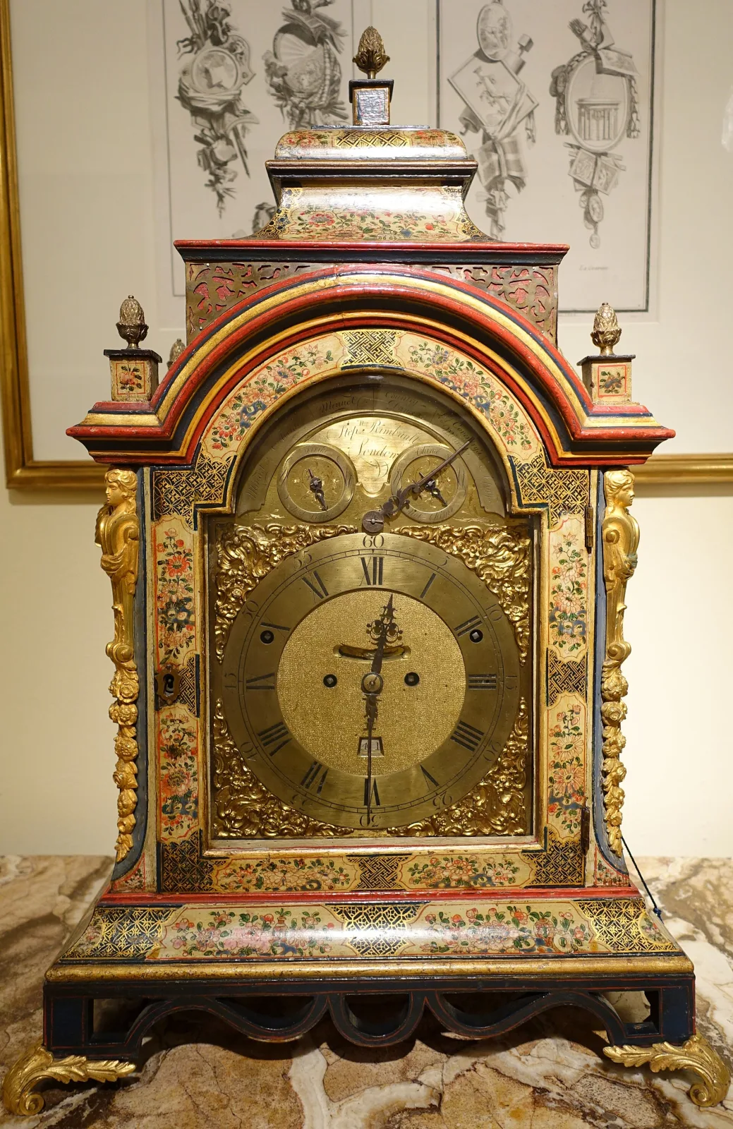 Ornate table clock in a red and gilt case with an arched top, a brass dial with subsidiary dials, and gilt figures and mounts against a patterned wall