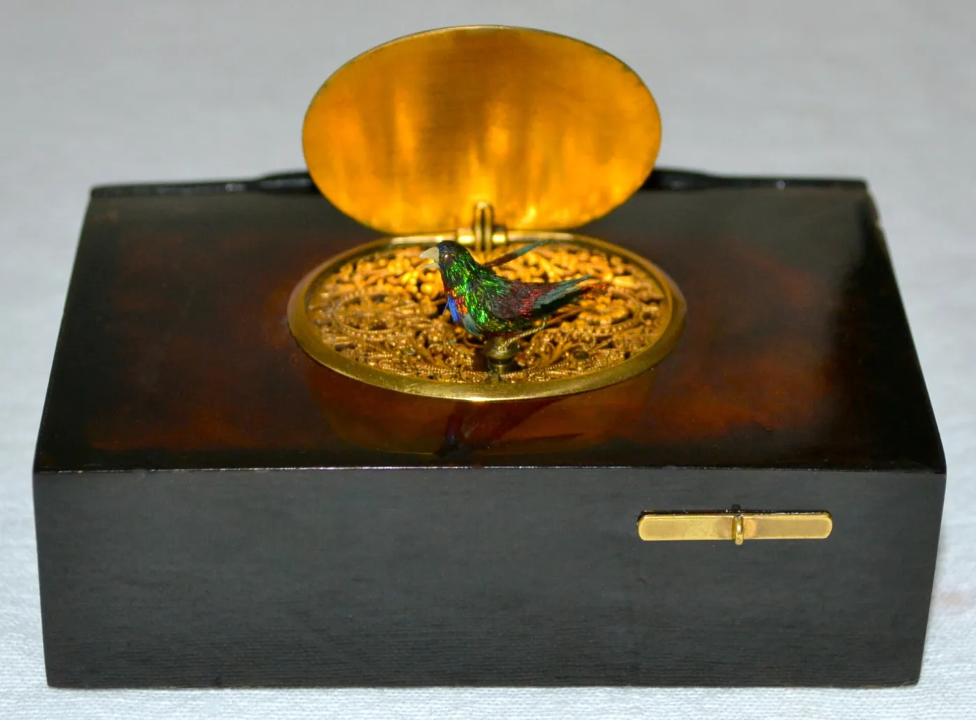 Dark rectangular box with its oval lid sprung open, revealing a pierced gilt grille and a small brightly feathered bird rising from the opening
