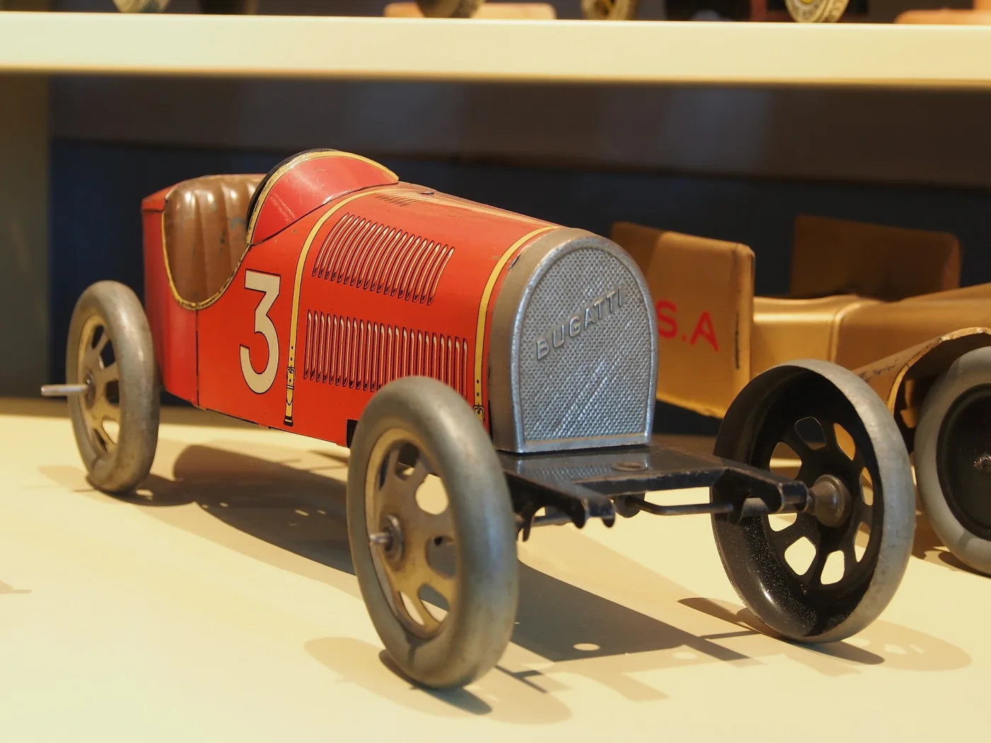 Pressed and lithographed tin toy racing car in red and cream with the number 3 on its side, spoked wheels and a visible winding key, standing on a shelf