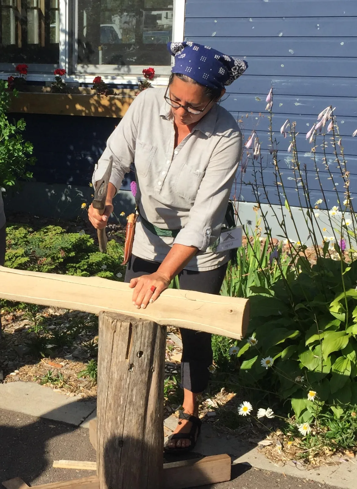 Woman in a headscarf standing outdoors and striking a long black ash log with a mallet, splitting pale fibrous layers away along its length