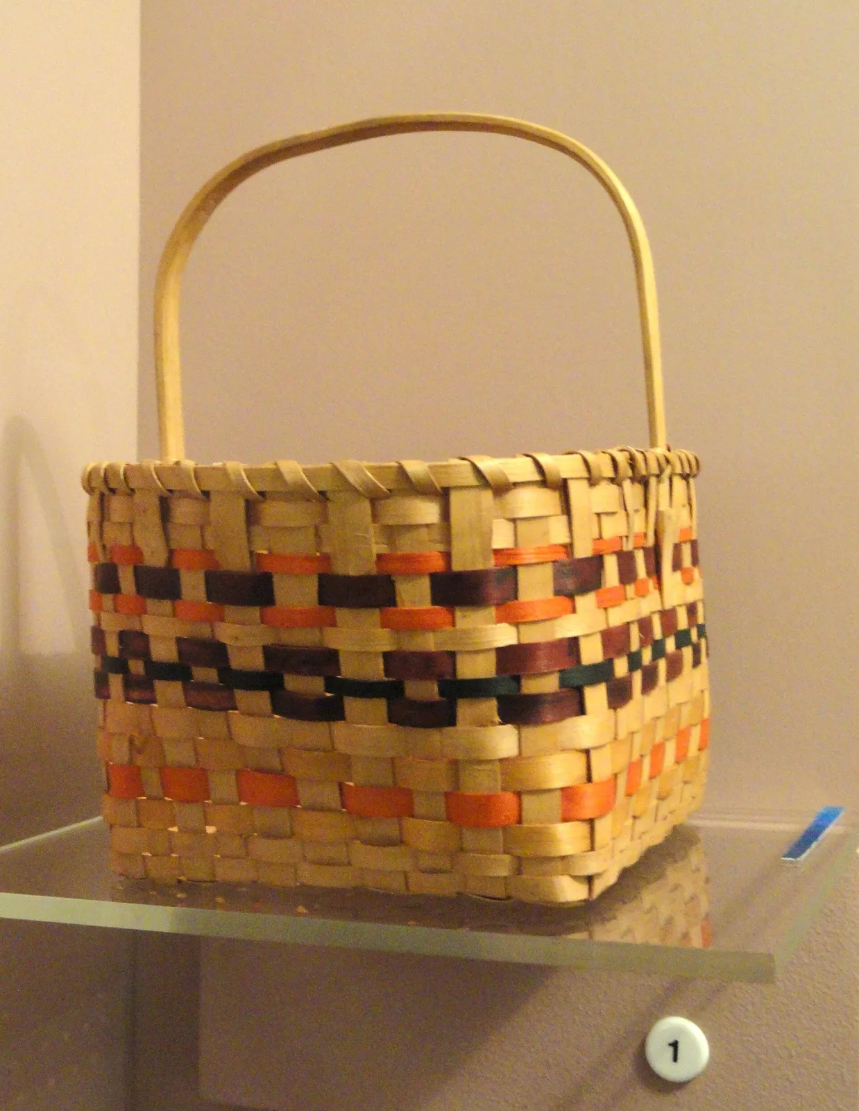 Rectangular basket woven from flat wooden splints in bands of natural, orange and black, with a wide bentwood handle arching over the top