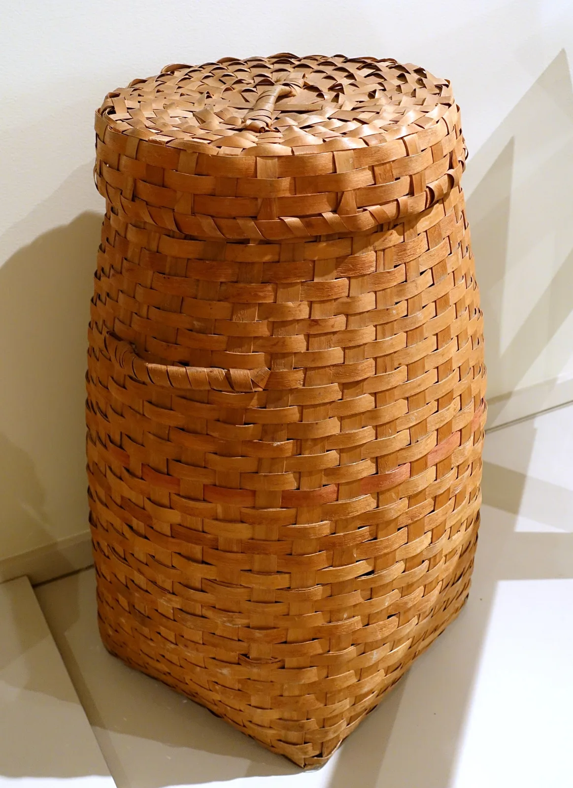 Tall cylindrical splint basket with a close-fitting domed cover, woven from broad pale wooden strips that darken to honey at the shoulders