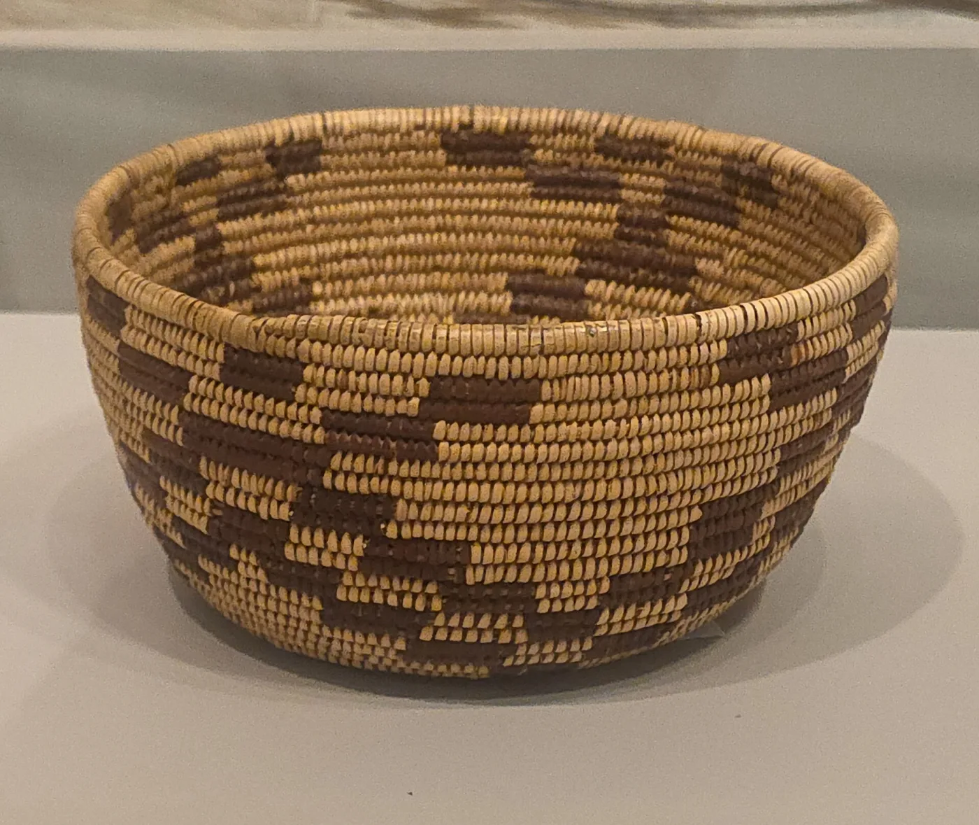 Coiled basket with a flaring rim, decorated with a band of stepped triangular figures in dark fibre against a pale ground, standing on a reflective surface