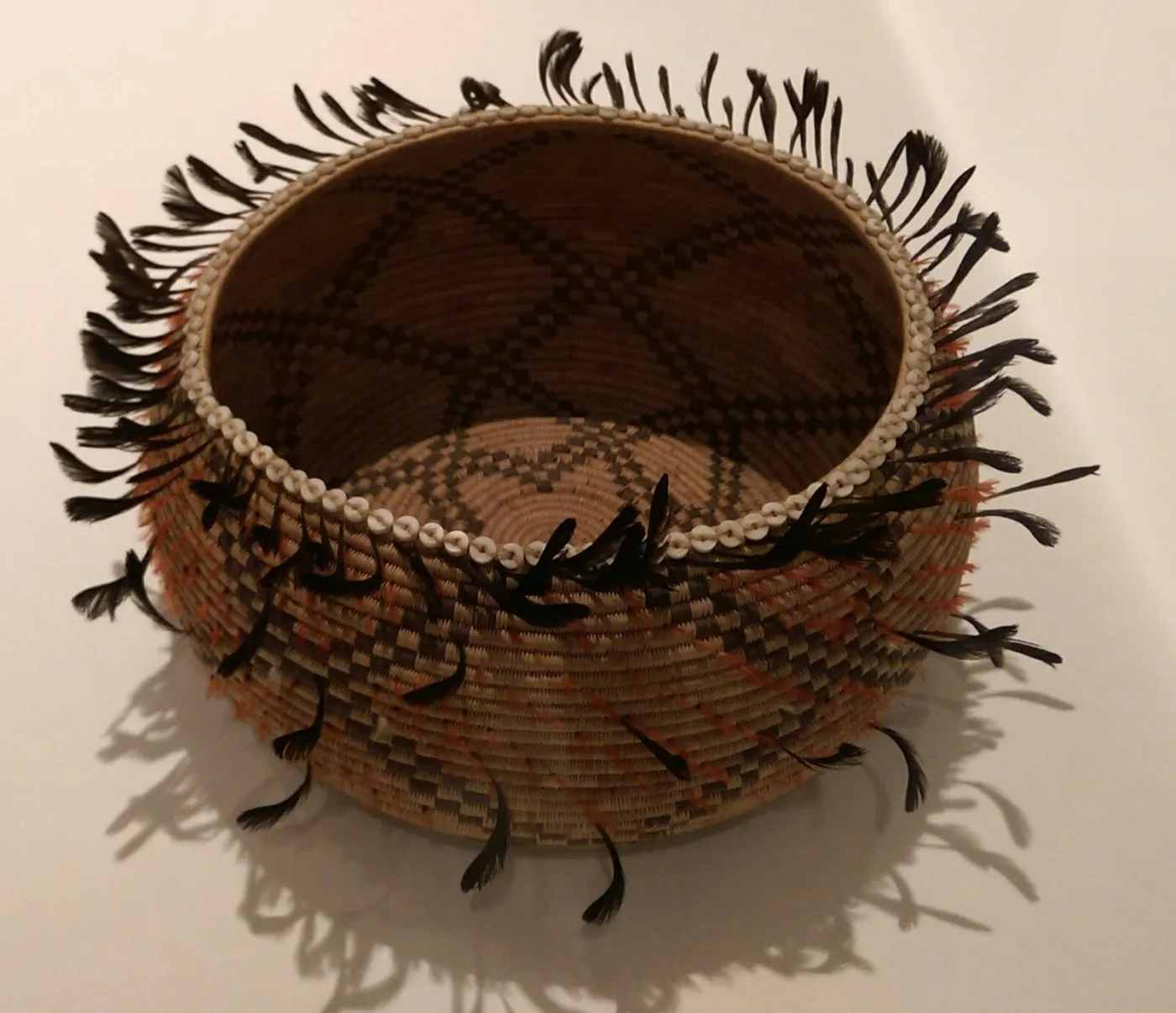 Shallow coiled basket whose surface is entirely covered in bright red and black feathers, with rows of white shell beads and long black quail plumes projecting from the rim