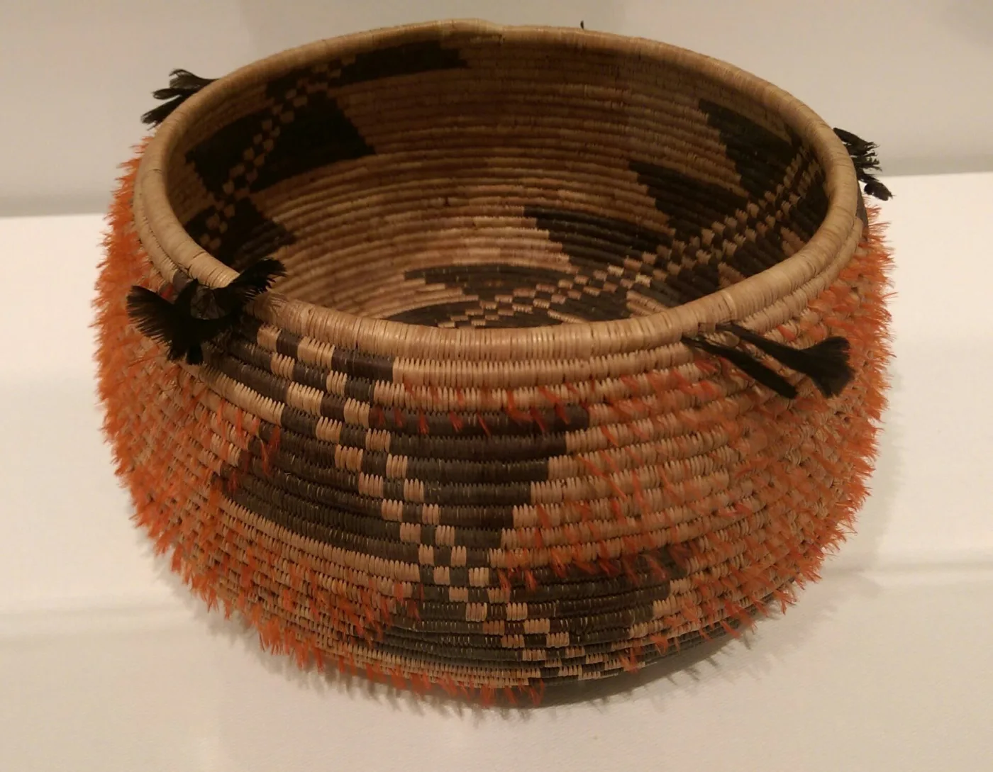 Coiled basket with a stepped black and orange geometric pattern, its rim fringed with short black quail crest feathers