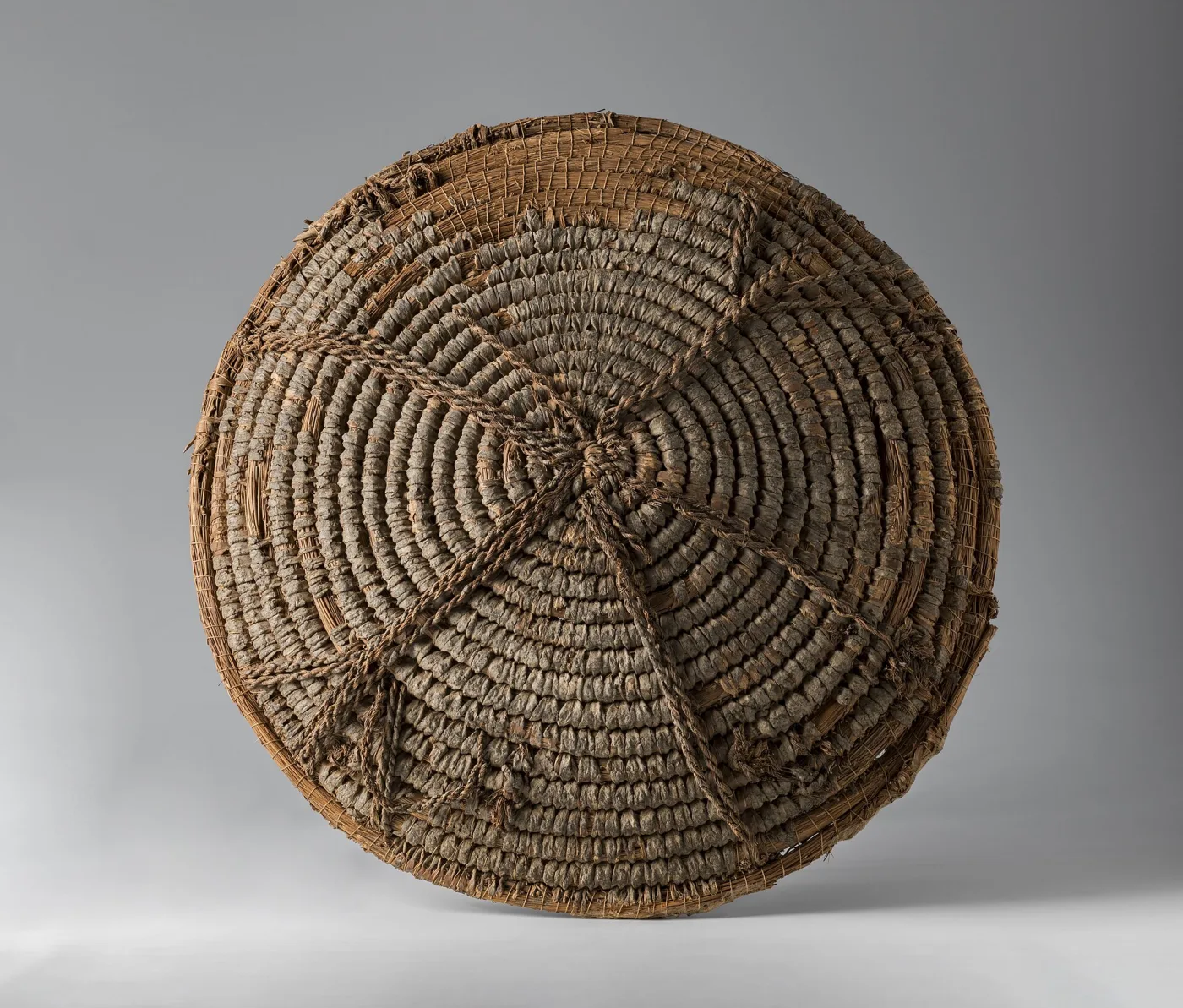 Round basket of pale vegetable fibre seen from above, its base built from a continuous spiral of bundled fibre stitched together, with a radiating pattern of darker stitching