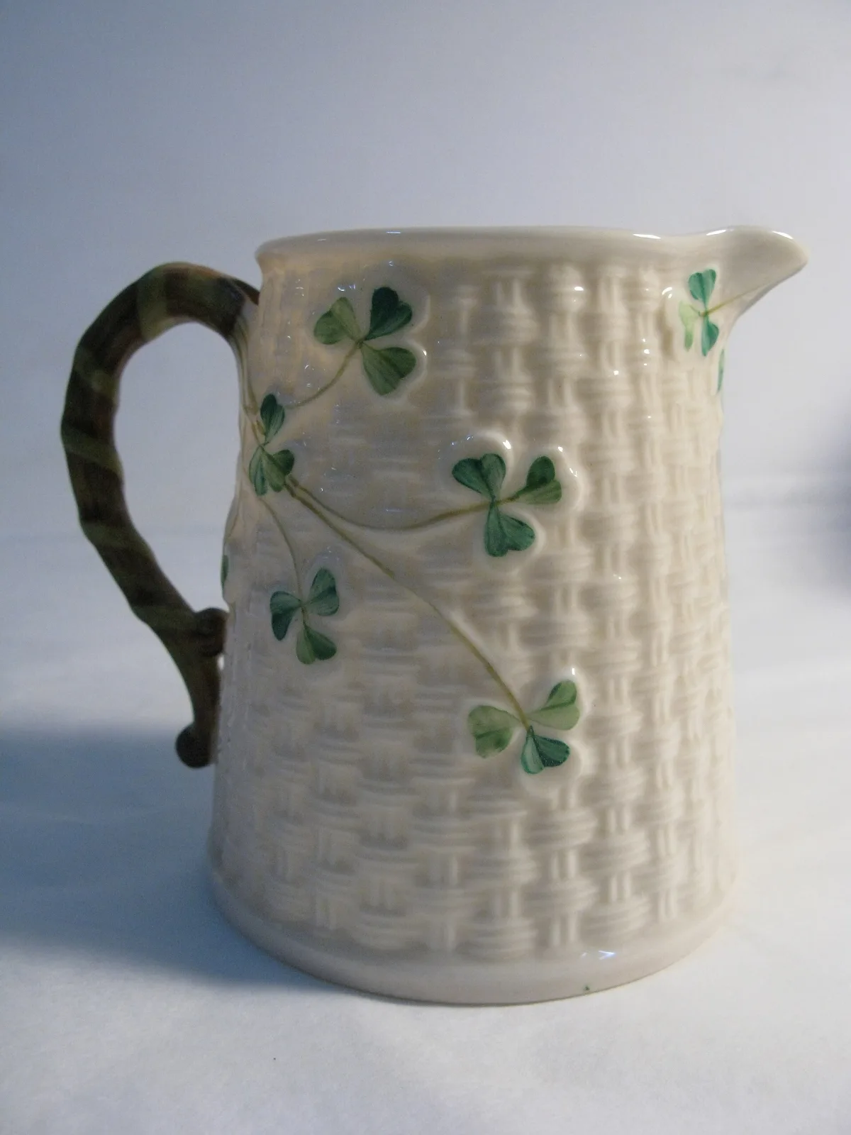 Small straight-sided porcelain milk jug with a moulded basket-weave ground, a twig handle and applied green shamrocks on the body