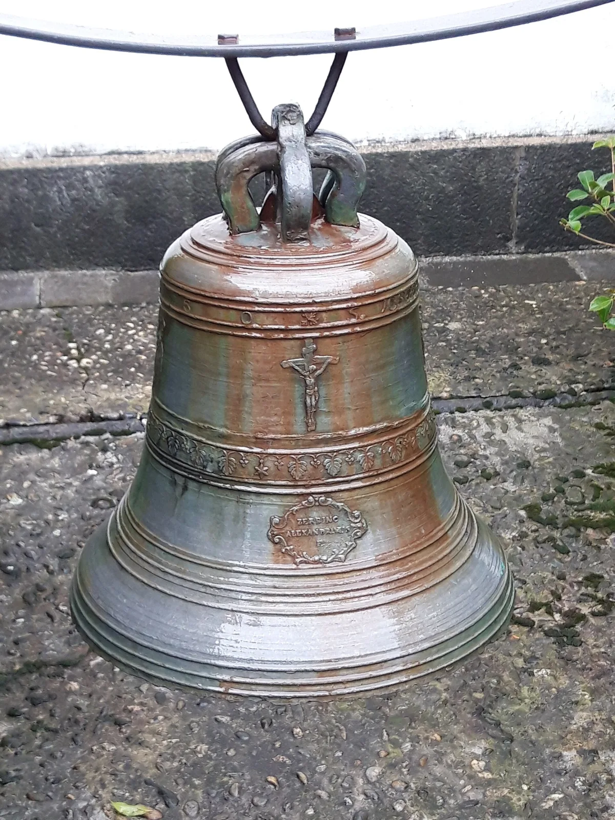 Bronze bell standing on a paved terrace, its surface streaked with green and brown patina, cast in relief with a crucifix and a small roundel on the waist