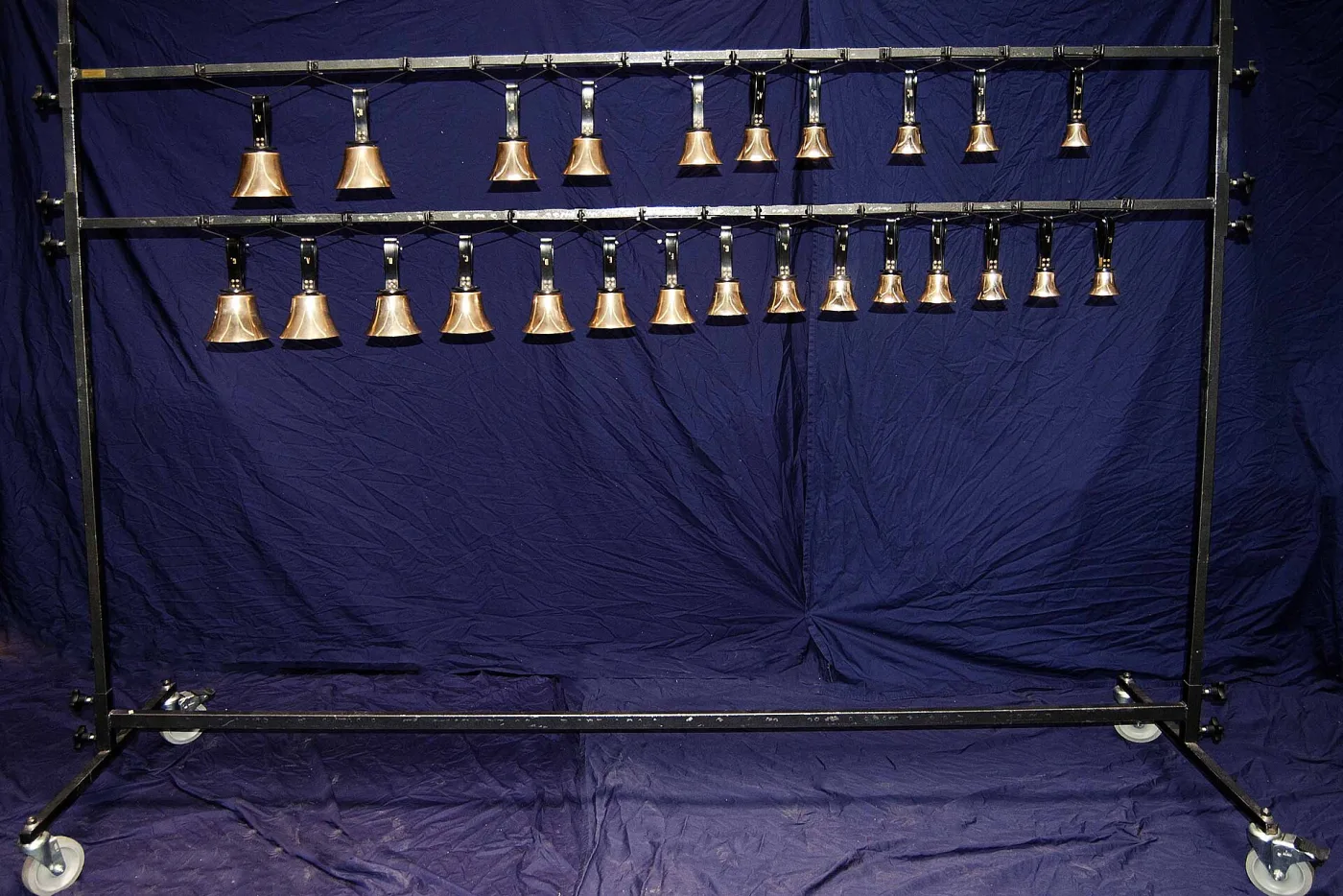 Row of about twenty polished brass hand bells of graduated sizes hanging in order along a metal stand against a dark blue cloth
