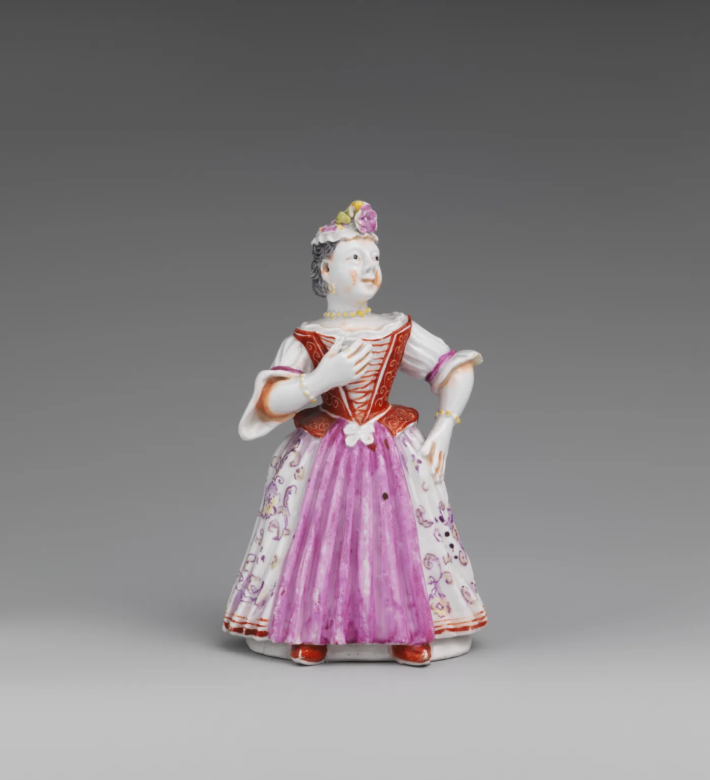 Porcelain table bell modelled as a standing woman in a pink and white dress with a laced bodice and flowered apron, her skirt forming the body of the bell