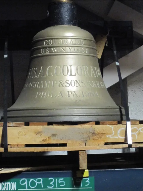 Ship's bell in a storage crate, its shoulder cast with lettering reading COLORADO, U.S. NAVY YARD, and WM CRAMP AND SONS, PHILA PA
