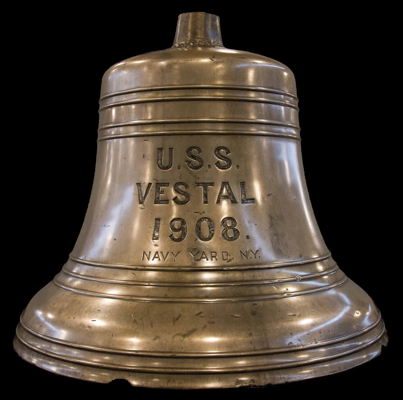 Large polished brass ship's bell photographed against black, its shoulder cast with raised lettering reading U.S.S. VESTAL 1908 NAVY YARD N.Y., with three moulding rings above and a flared lip below