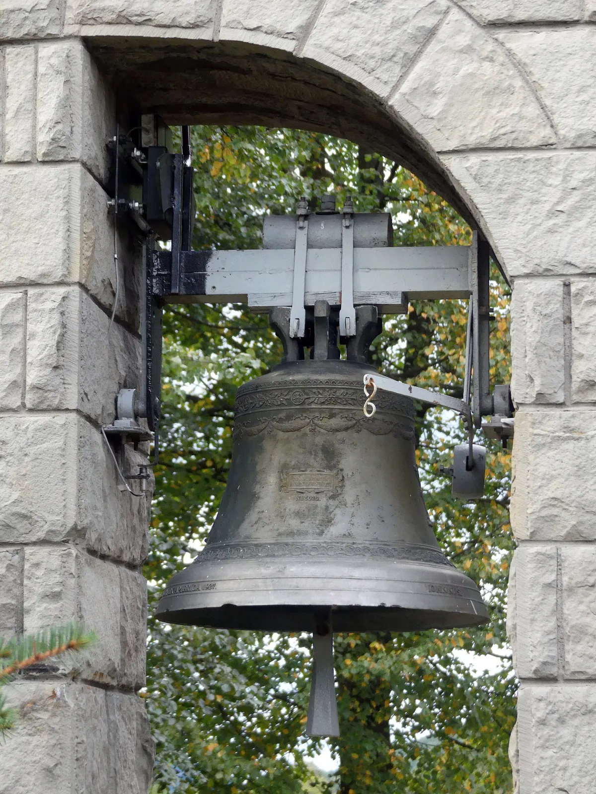 Bronze bell hanging from a wooden headstock inside a stone belfry arch, seen against daylight, with a Latin inscription visible around its shoulder