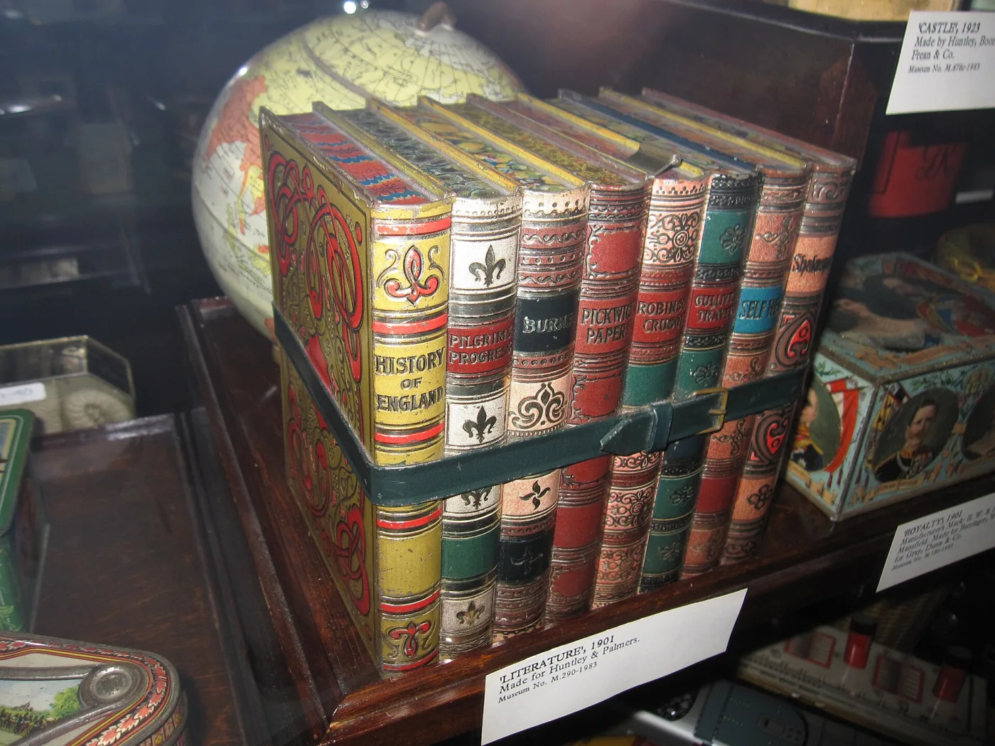 A biscuit tin in the form of eight leather-bound books held by a strap, displayed in a museum case beside a printed label reading Literature, 1901