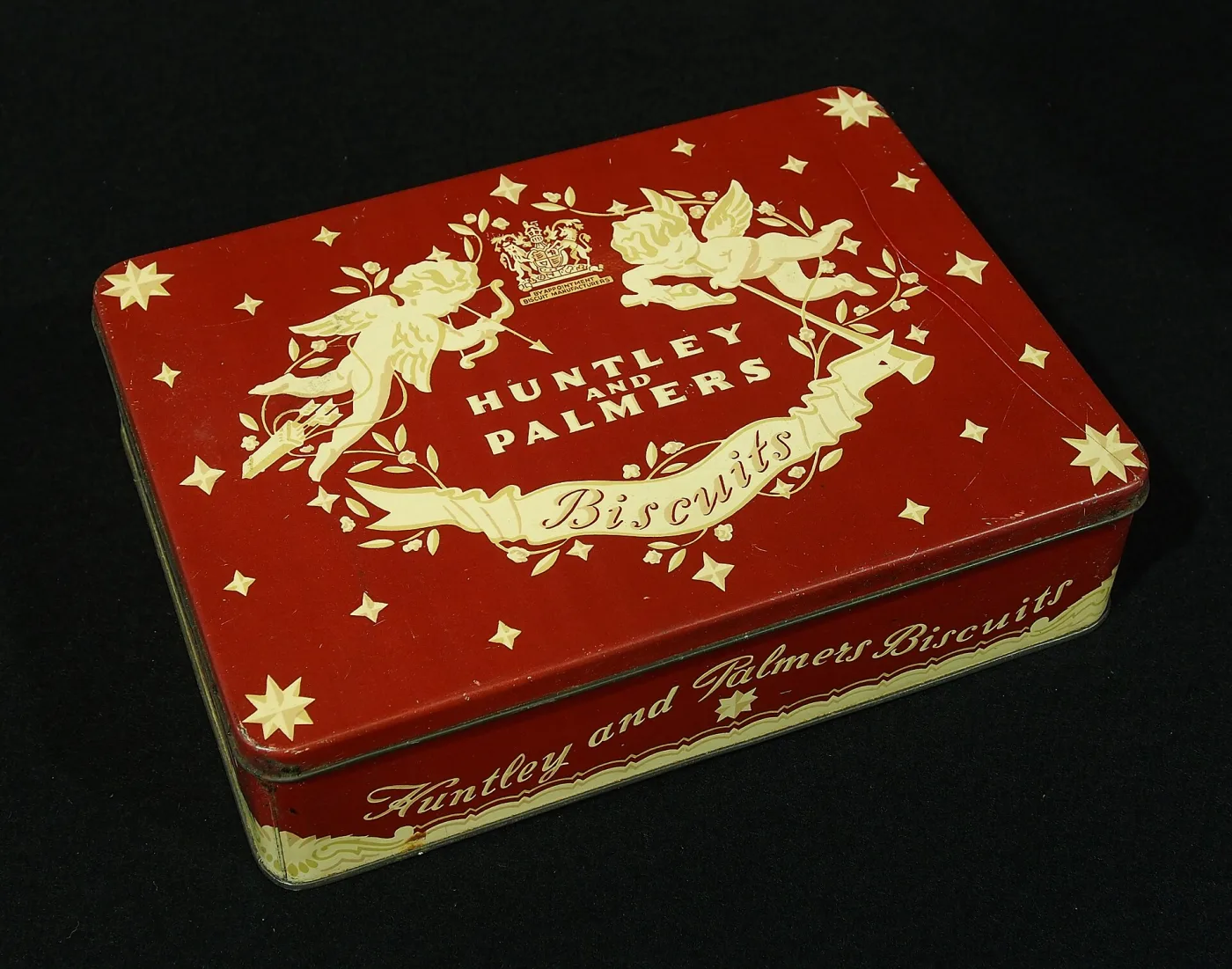 A rectangular red biscuit tin lithographed in cream with two cherubs, stars, a royal coat of arms and the words Huntley and Palmers Biscuits