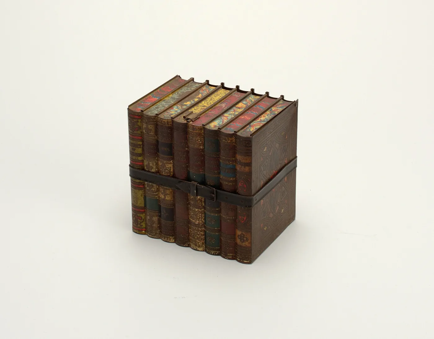 A biscuit tin in the form of eight bound books secured by a strap and buckle, the hinged lid printed as marbled page edges, photographed against a plain pale ground