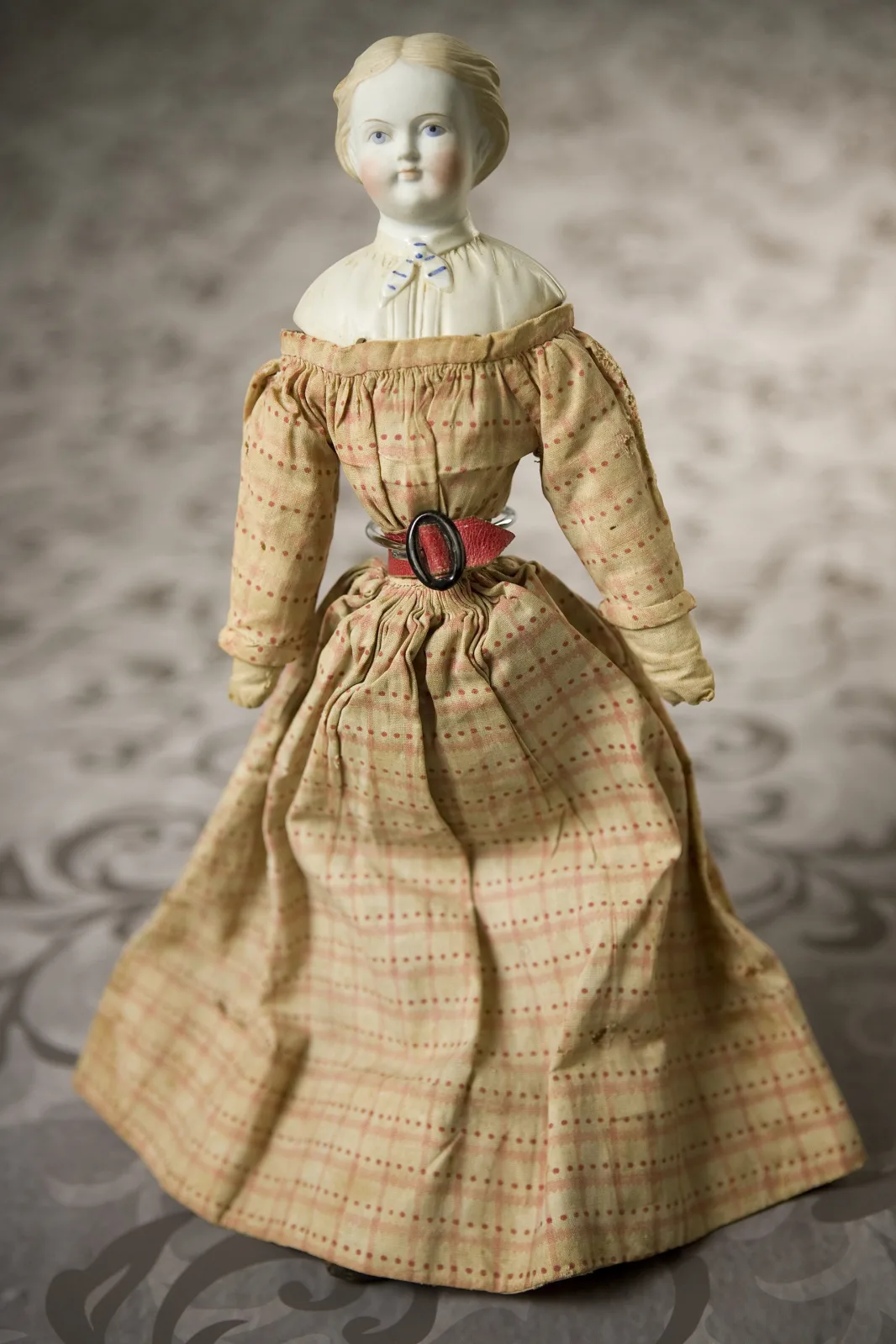 Doll with a bisque shoulder head, moulded and painted blond hair, and a soft cloth body, wearing a printed cotton dress with a wide red leather belt