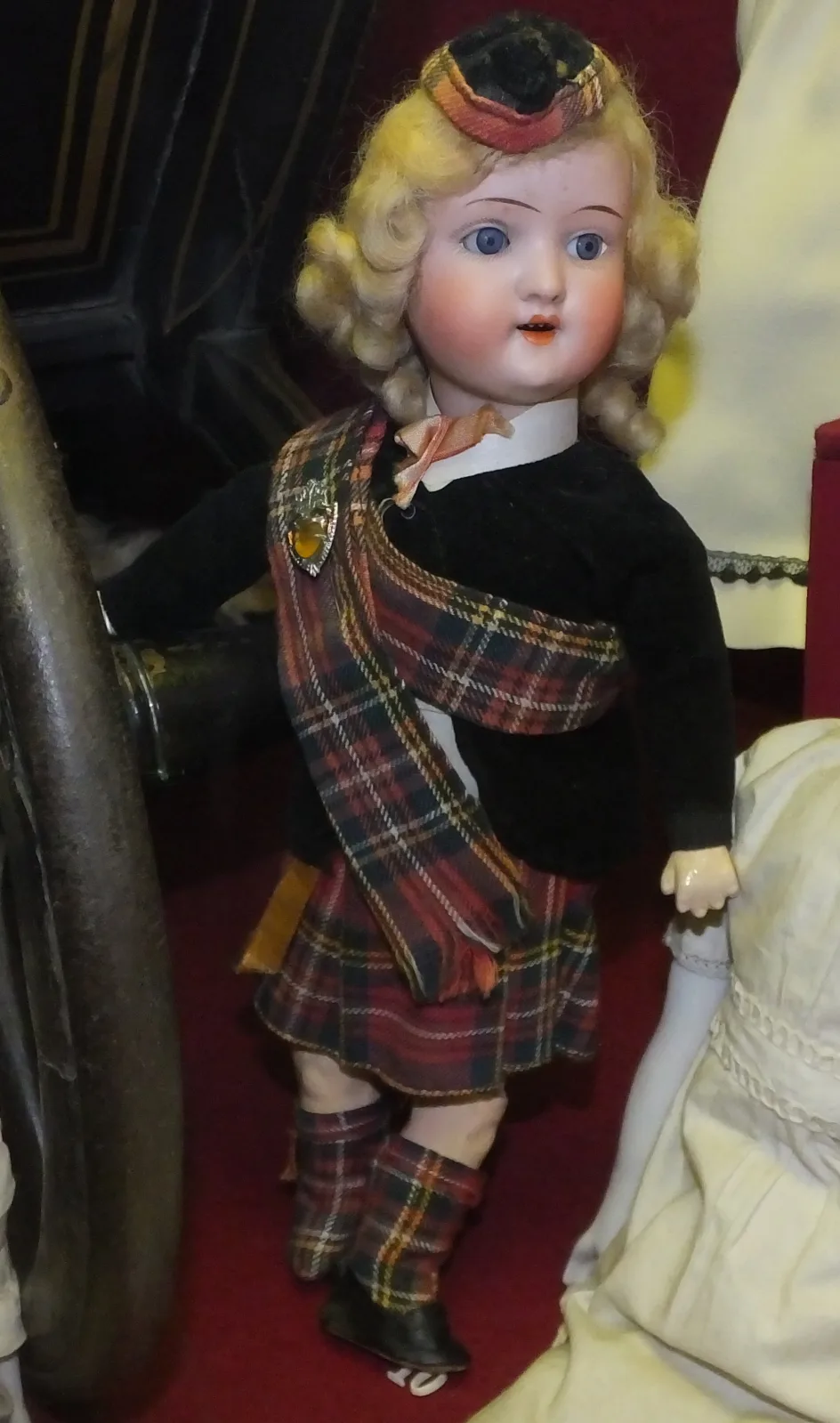 Doll in Highland dress with a tartan sash and kilt, a feathered bonnet and a bisque head with glass eyes and an open mouth, displayed in a museum case
