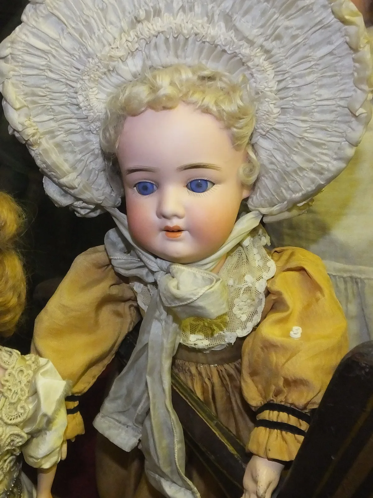 Close view of a doll's bisque head in a wide frilled bonnet, showing blue glass eyes, painted lashes and brows, a slightly open mouth and a blonde mohair wig