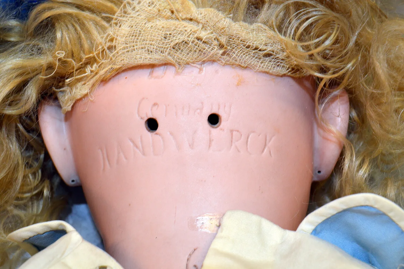 Back of a doll's head with the wig lifted away, showing the pale bisque crown incised with a maker's name and the word Germany, and two small stringing holes
