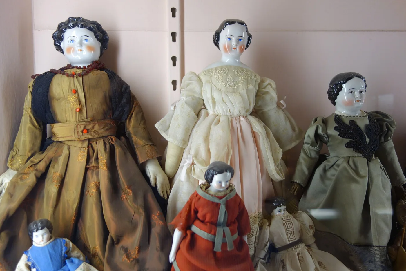 Museum shelf holding a row of dolls with glossy white china heads and painted black hair, dressed in period gowns, with smaller dolls in front