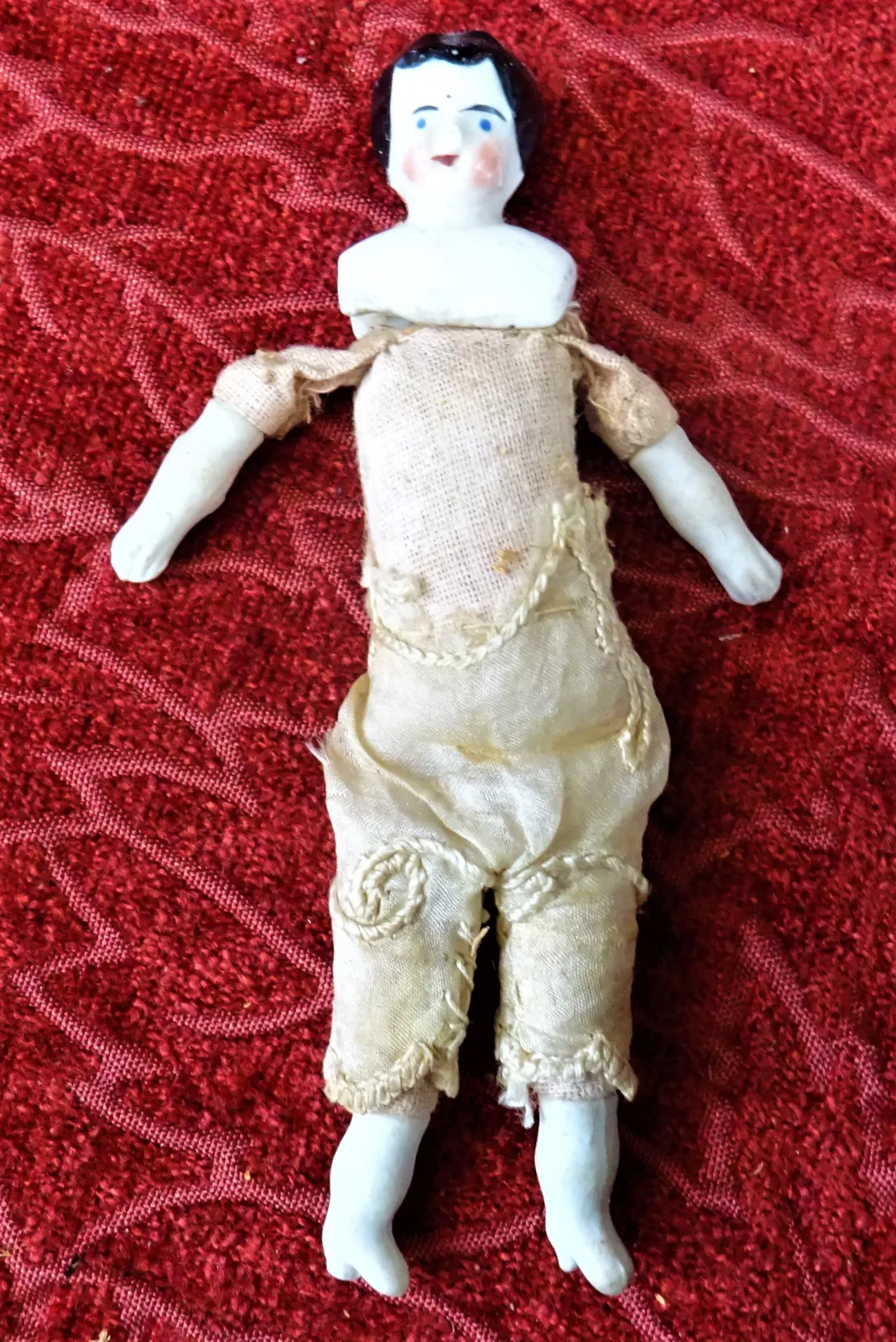 Small doll about ten centimetres long lying on red fabric, with a glazed china head and lower limbs and a plain stuffed cloth body and underclothes