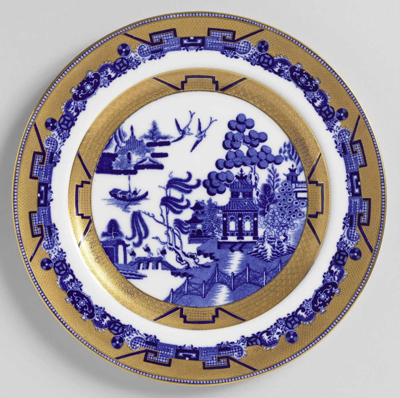 Gilded plate seen from directly above: a small blue Willow scene in a central medallion, ringed by a wide gold band carrying dark blue fretwork panels and floral reserves