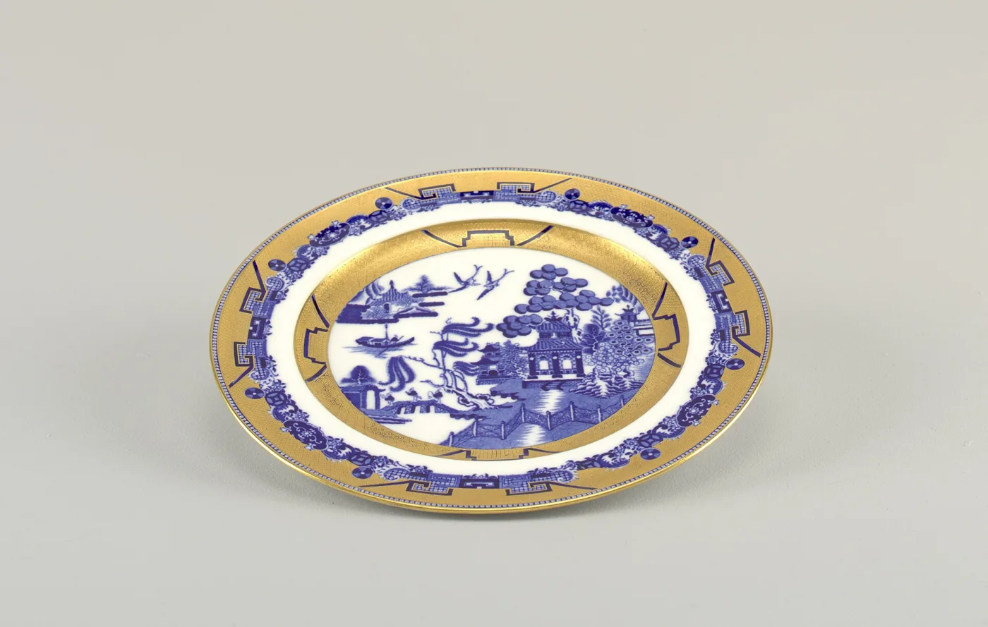 The same gilded Willow plate photographed at a low raking angle, the gold band catching the light across the rim