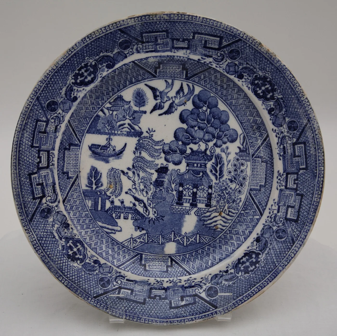 Blue and white transfer-printed plate on a stand, showing three small figures crossing a bridge, a pagoda among trees, a fence and a pair of birds overhead