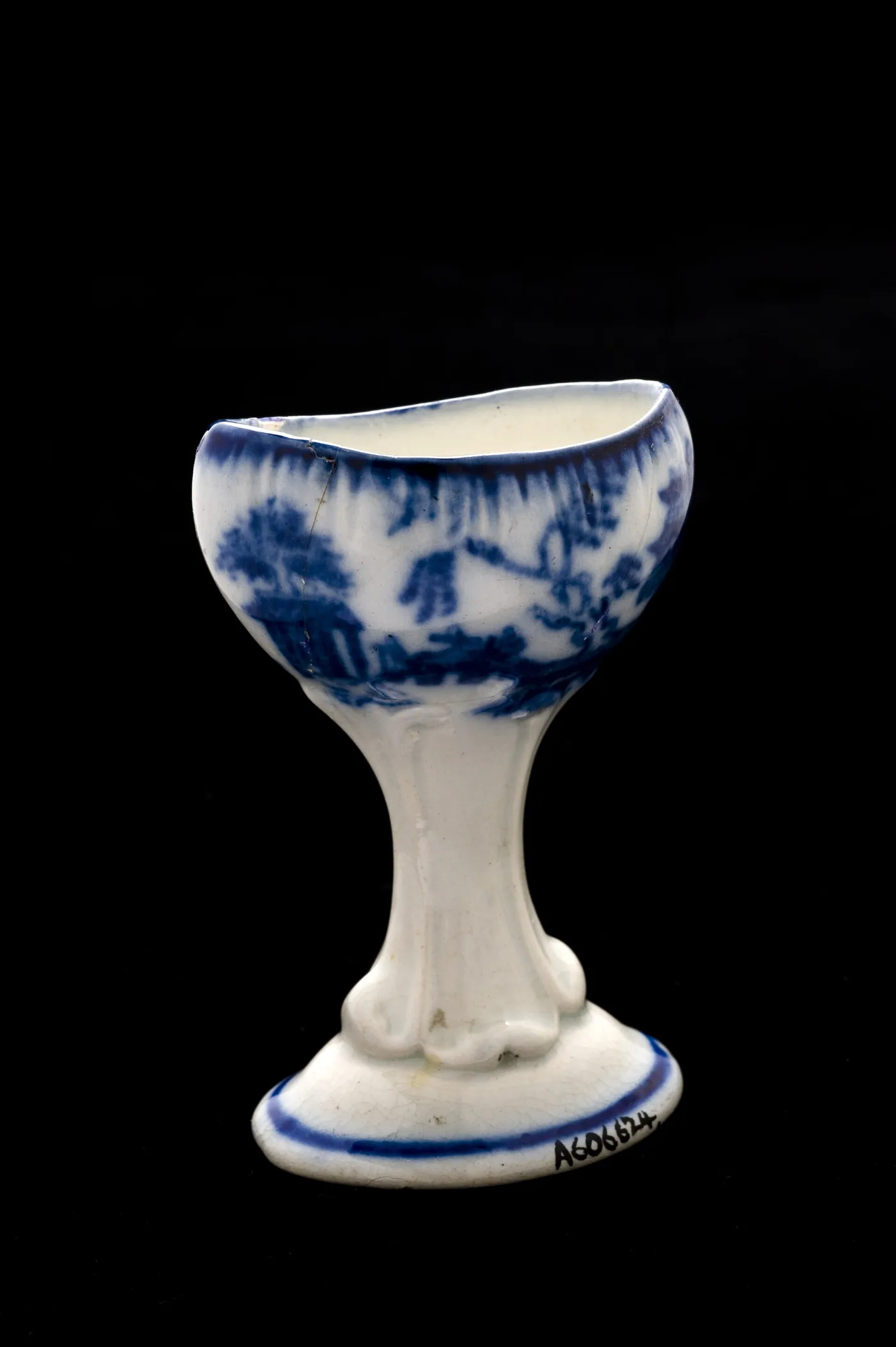 Small footed eye bath on a moulded stem, its oval bowl printed in blurred blue with a willow scene that fades into the white glaze, photographed against black