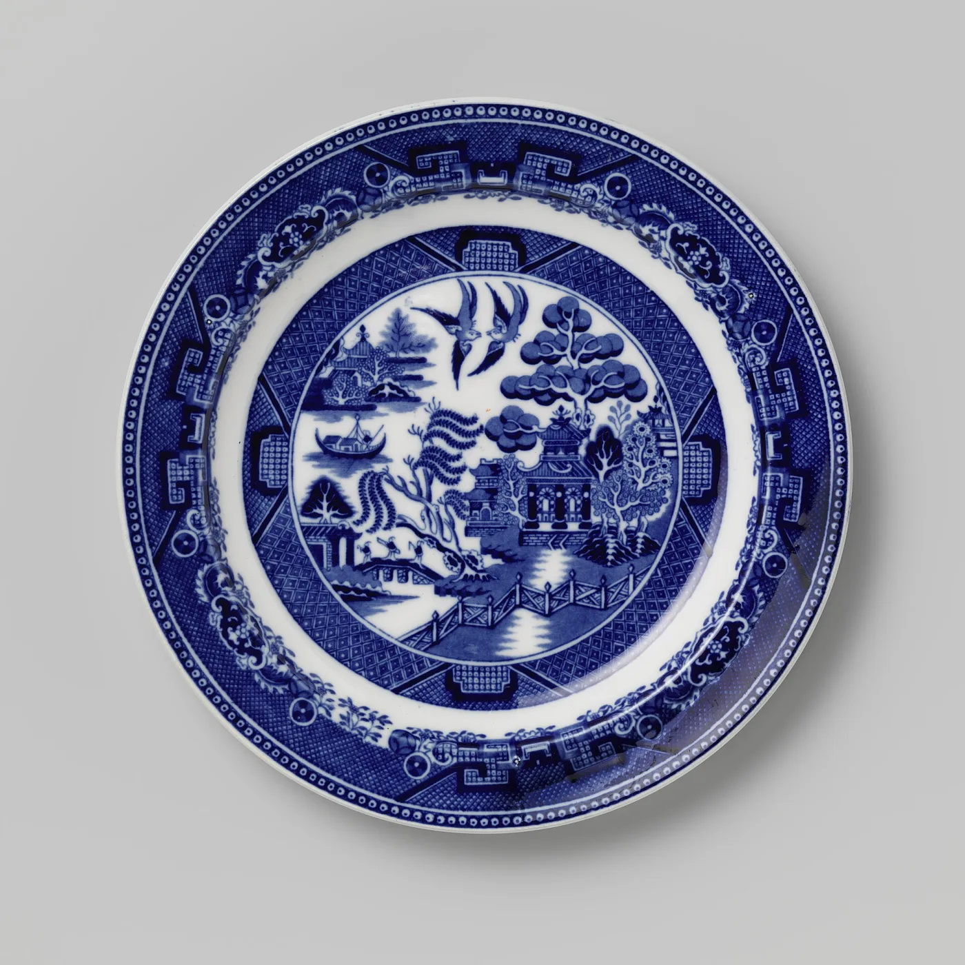 Round blue and white plate printed with a willow tree, a many-roofed teahouse, a bridge with small figures, a boat and two birds, inside a wide geometric border
