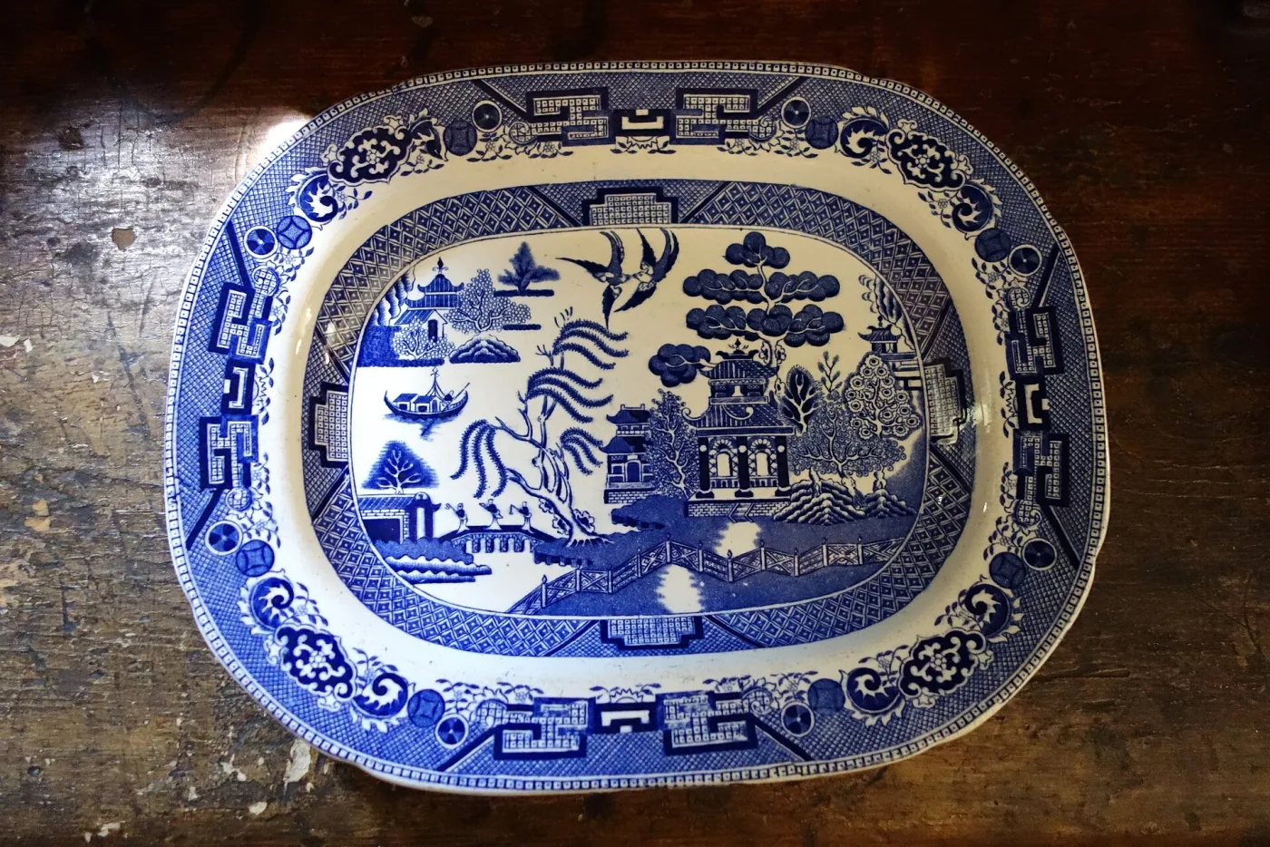 Large oval blue and white Willow platter lying on a worn wooden table, its rim border printed separately from the central scene