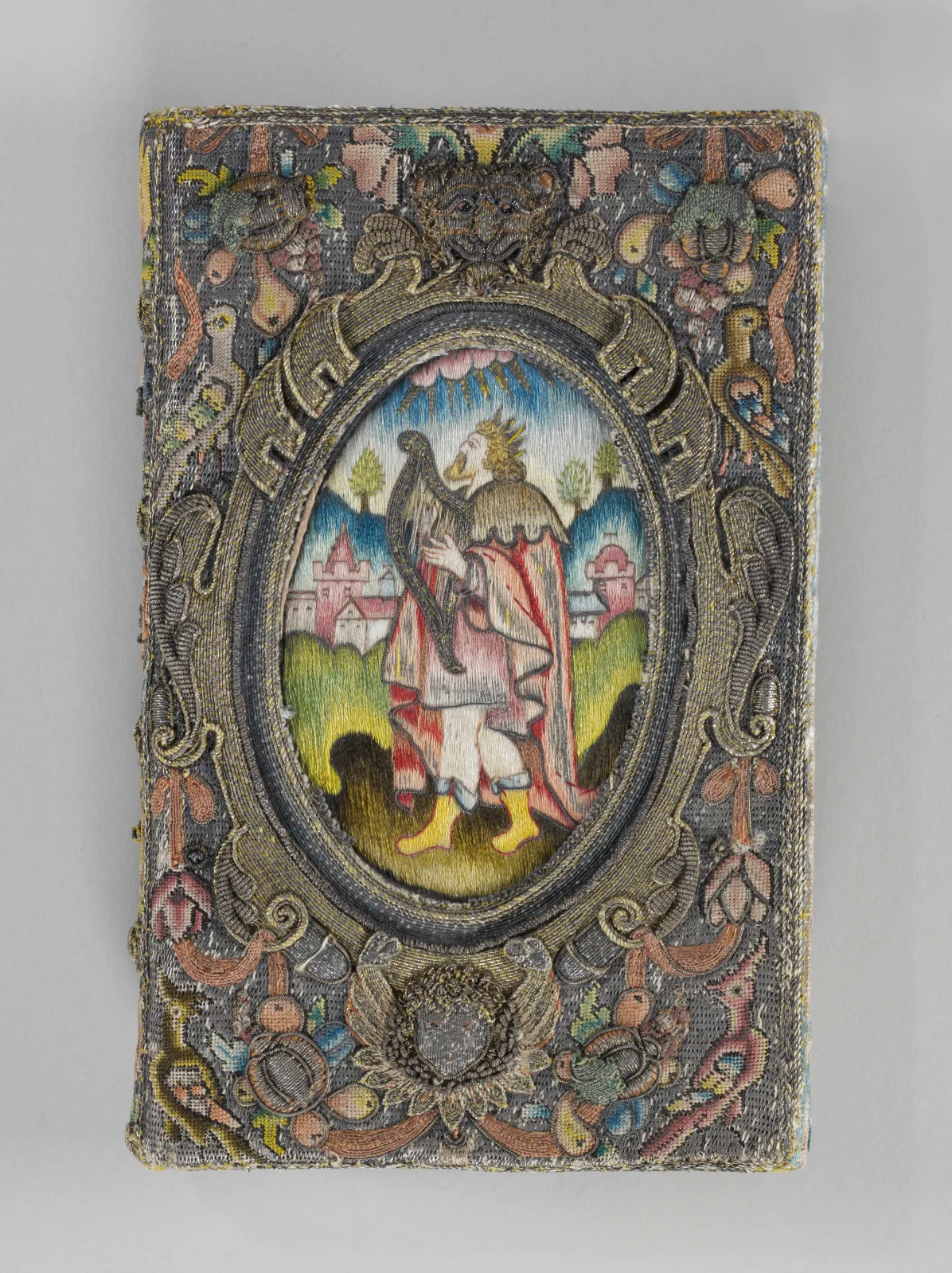 Book cover embroidered in coloured silks and metal thread, with a central oval scene of a figure in a landscape surrounded by dense floral and foliate embroidery worked in silver and gilt