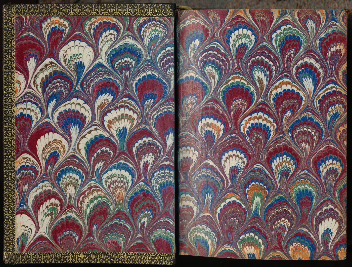 Open book showing both endpapers marbled in a dense repeating pattern of arched peacock-tail shapes in red, blue and cream, with a gilt-decorated edge visible at the left