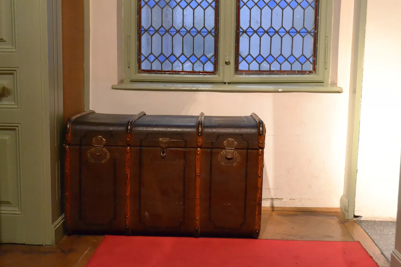 Leather-covered dome-top trunk with three iron-bound straps running over the lid, standing on a red carpet in a museum room beside a leaded window