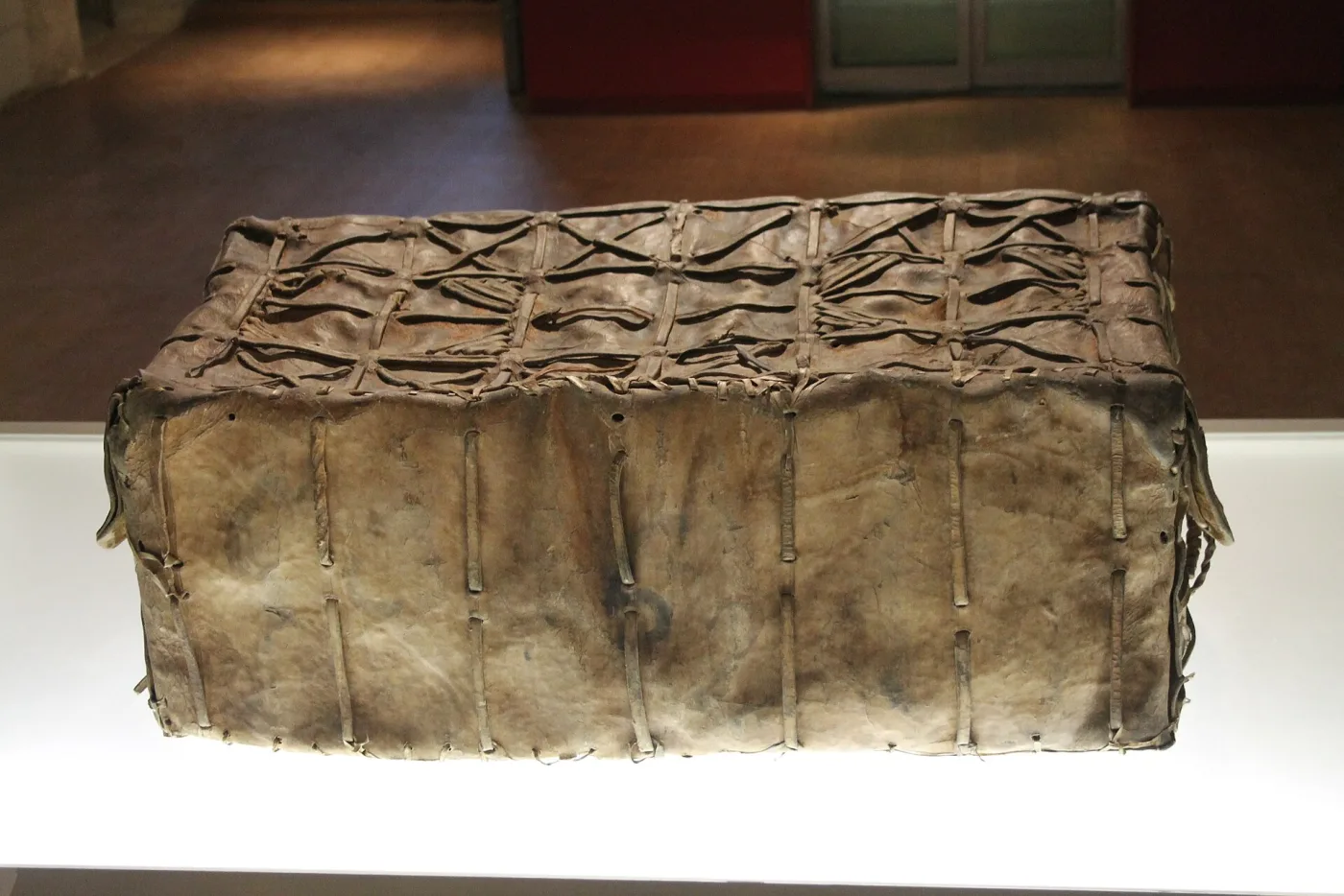 Low rectangular chest covered in pale hide laced together with leather thongs along its edges, displayed on a plain white plinth