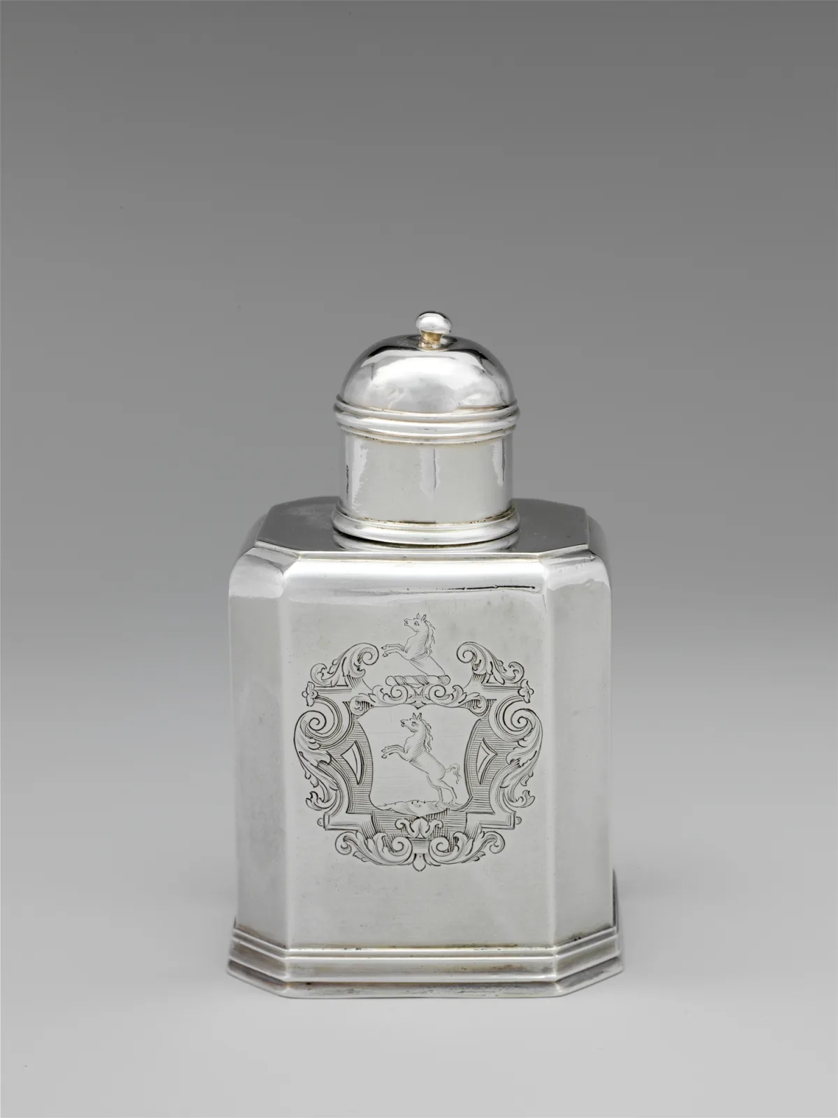 Plain silver tea caddy of canted rectangular form with a domed cover and small finial, engraved with an armorial cartouche on the front